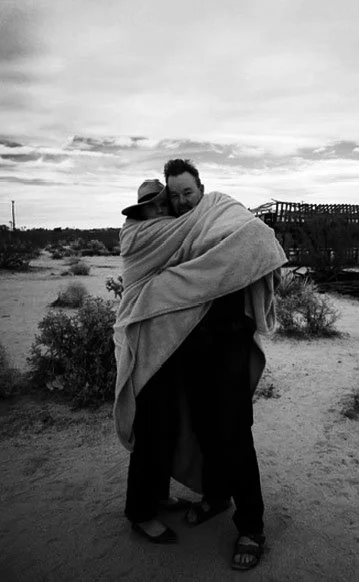 Two people hugging in a desert landscape with cloudy sky and distant structures.