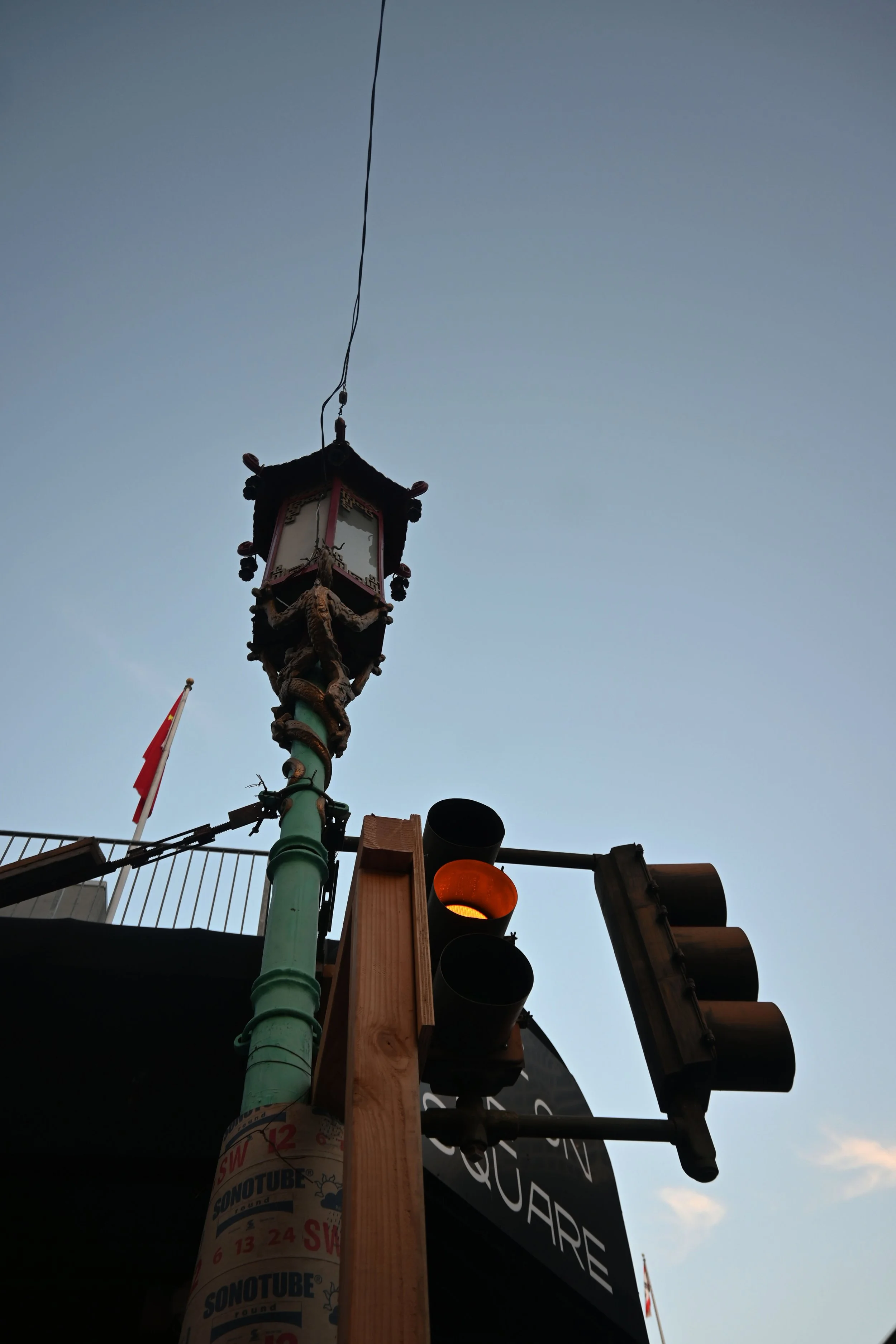 Close-up of a decorative street lamp with dragon motifs, a traffic light showing orange, and a wooden support structure against a clear sky at dusk.