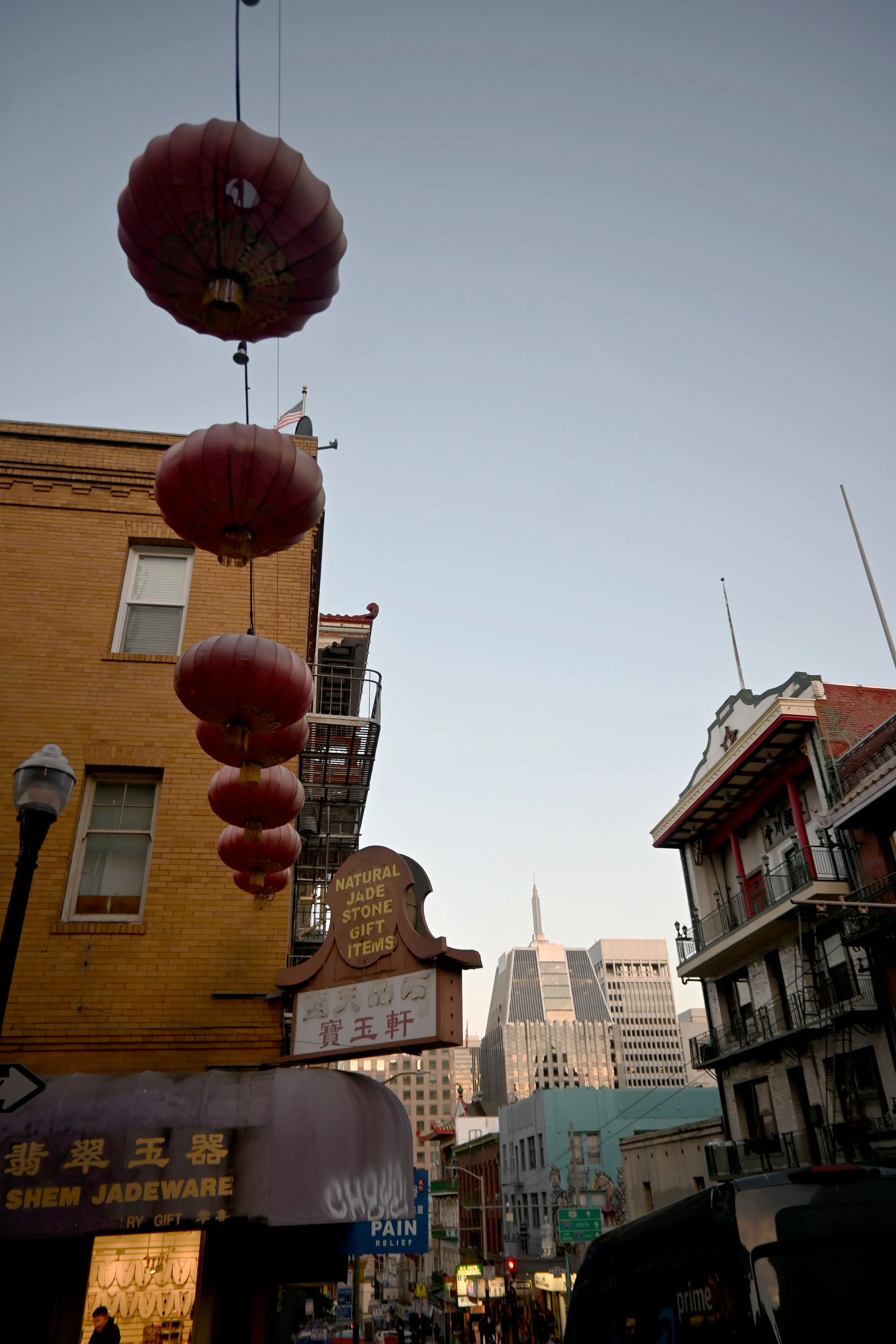 Street view in Chinatown with Chinese lanterns hanging, brick buildings, modern skyscrapers in the background, and signs in Chinese and English.