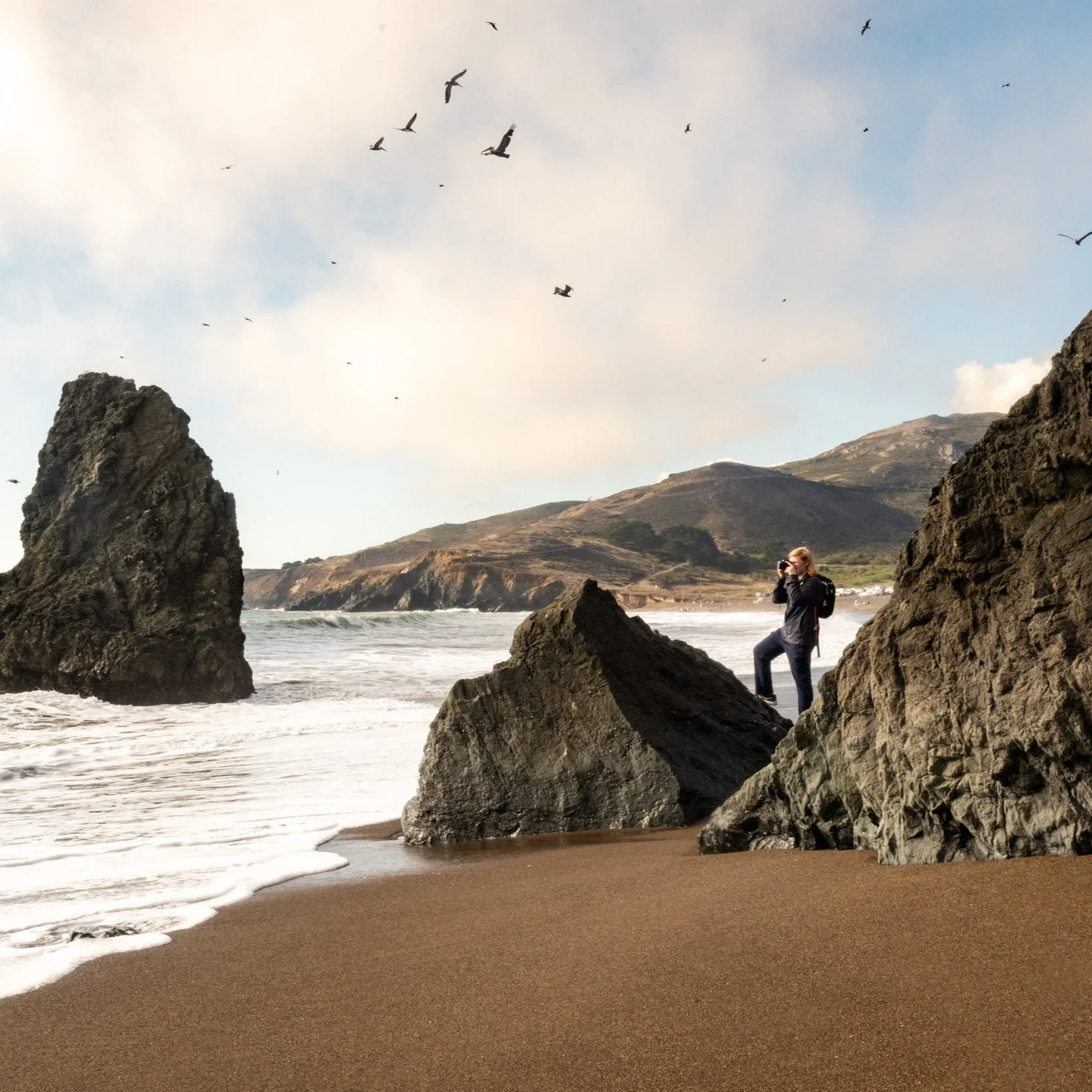 Person taking photos on a rocky beach with large rocks, ocean waves, and bird flock in the sky, with hills in the background.