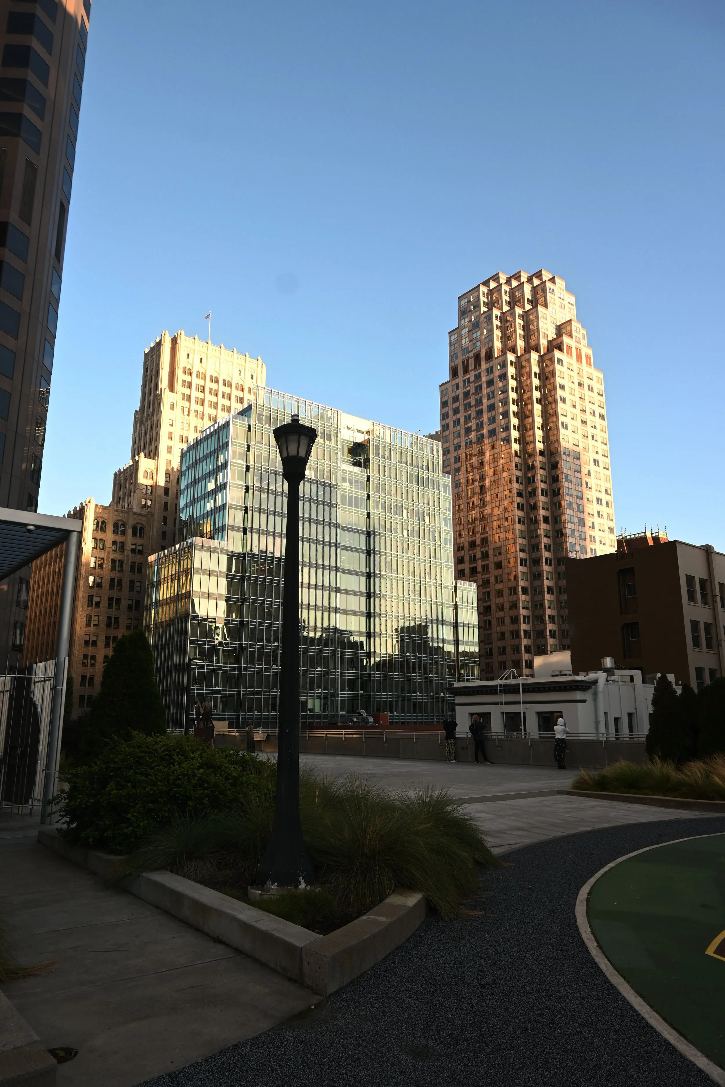 Cityscape with tall buildings, some with glass facades, under a clear blue sky, and a pathway with people near street lamps and greenery.