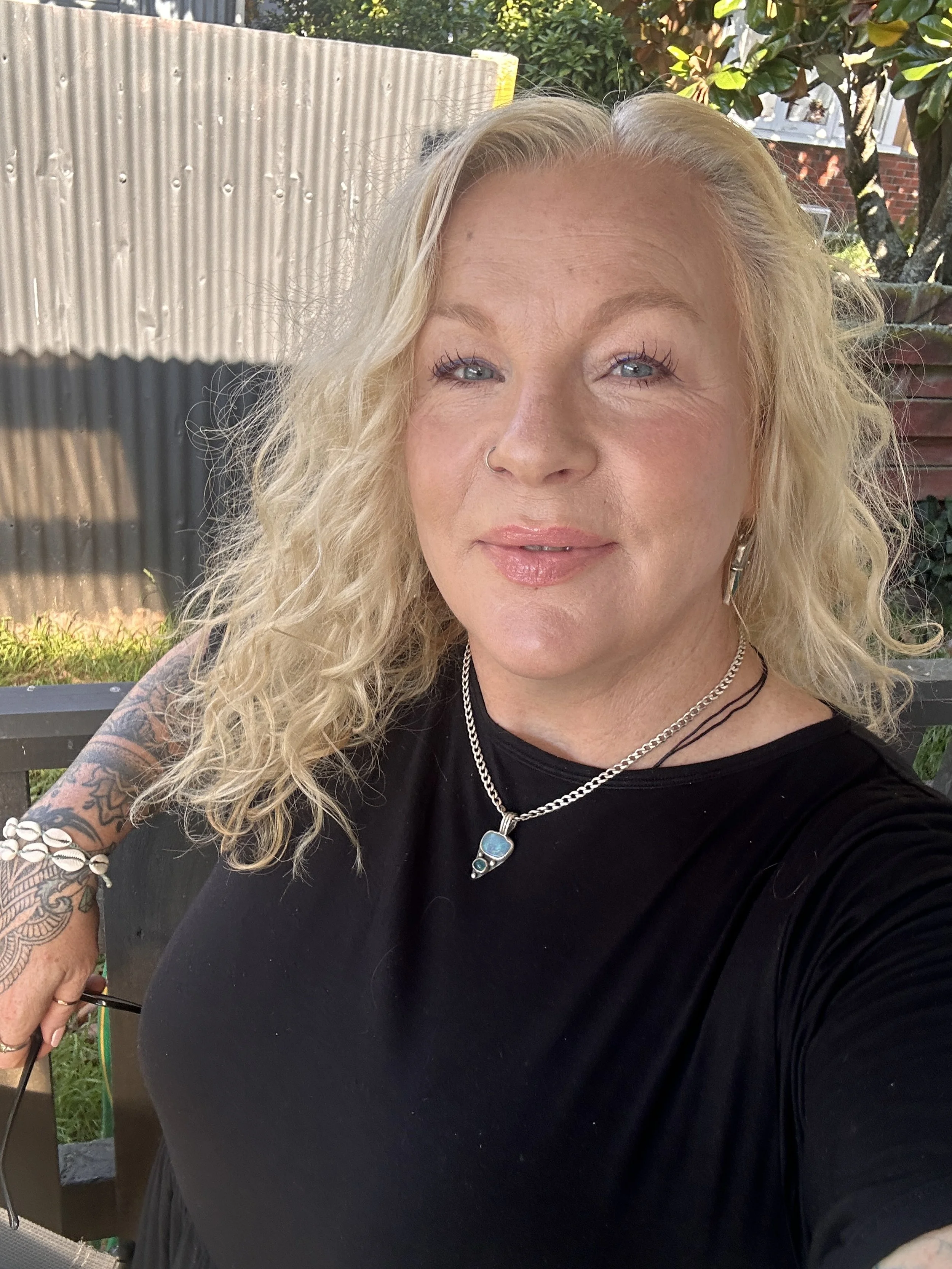 A woman with blonde curly hair taking a selfie outdoors. She is wearing a black top, jewelry, and has tattoos on her arm.