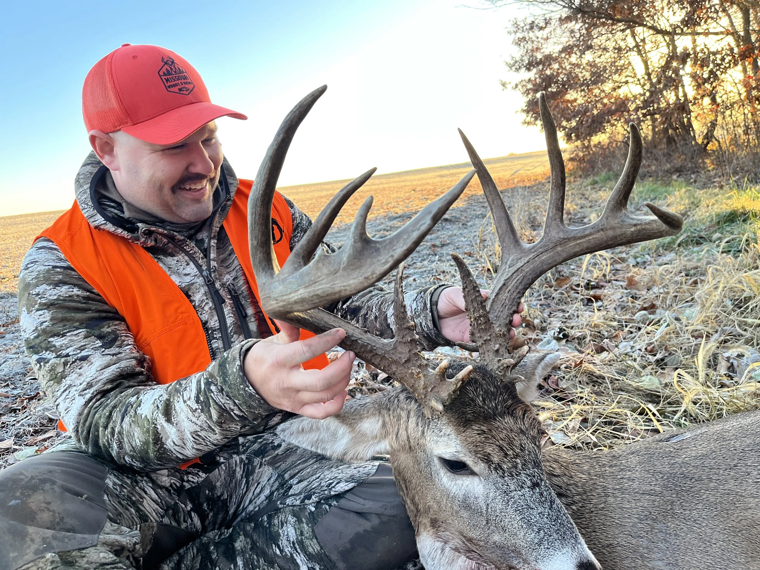 A man in camouflage and orange hunting gear holding a large deer with antlers, sitting outdoors during sunset.