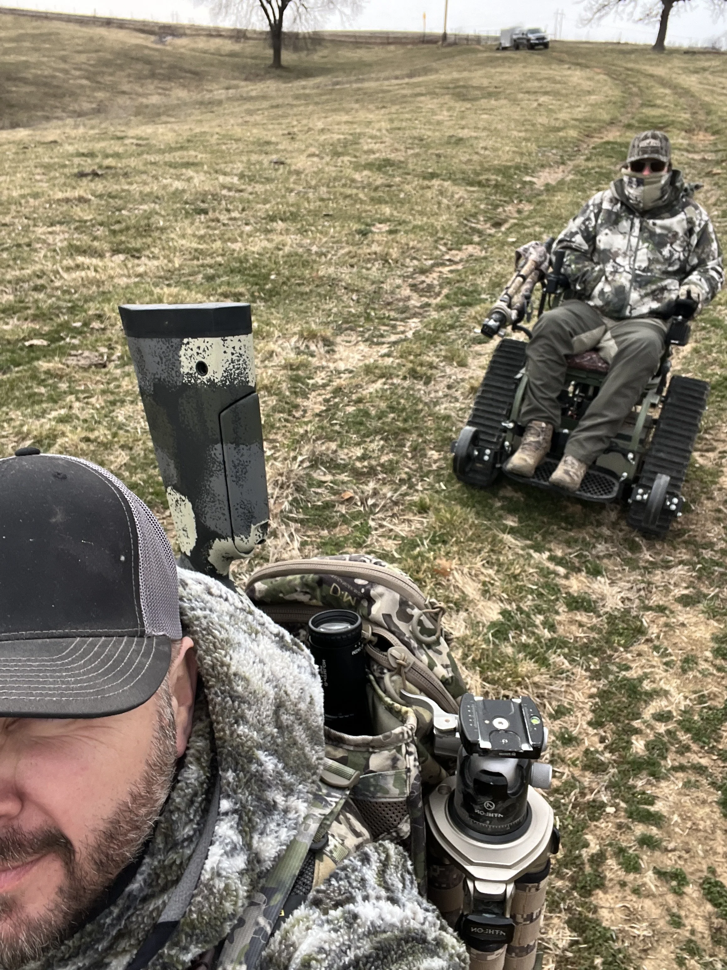 A person in camo gear taking a selfie outdoors near a motorized wheelchair with a person in camo gear sitting on it, in a vast open grassy area with trees in the background.