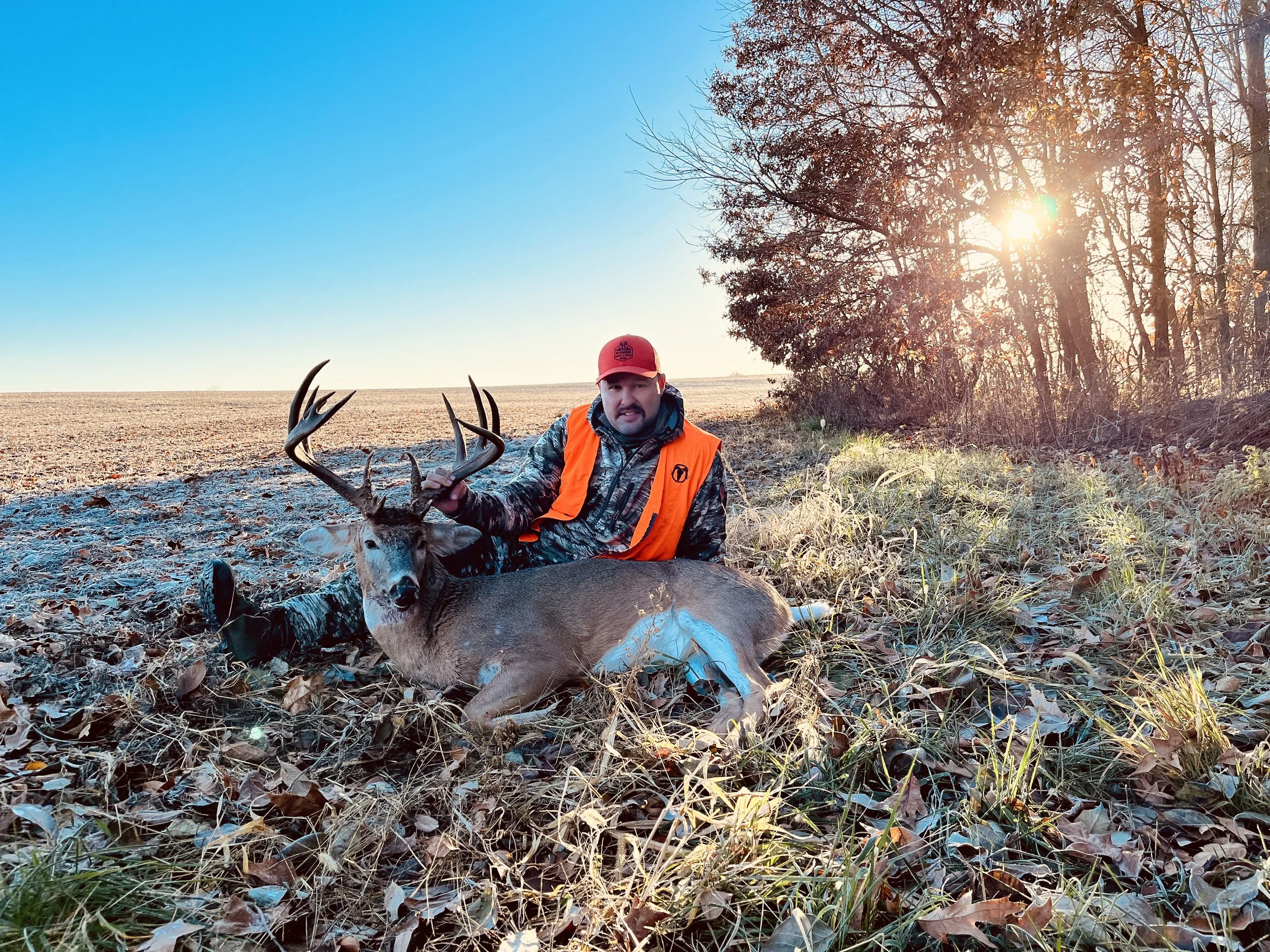 A man in a camouflage outfit, orange vest, and red cap poses with a large deer he has hunted, sitting on a frosty field with trees and a low sun in the background.