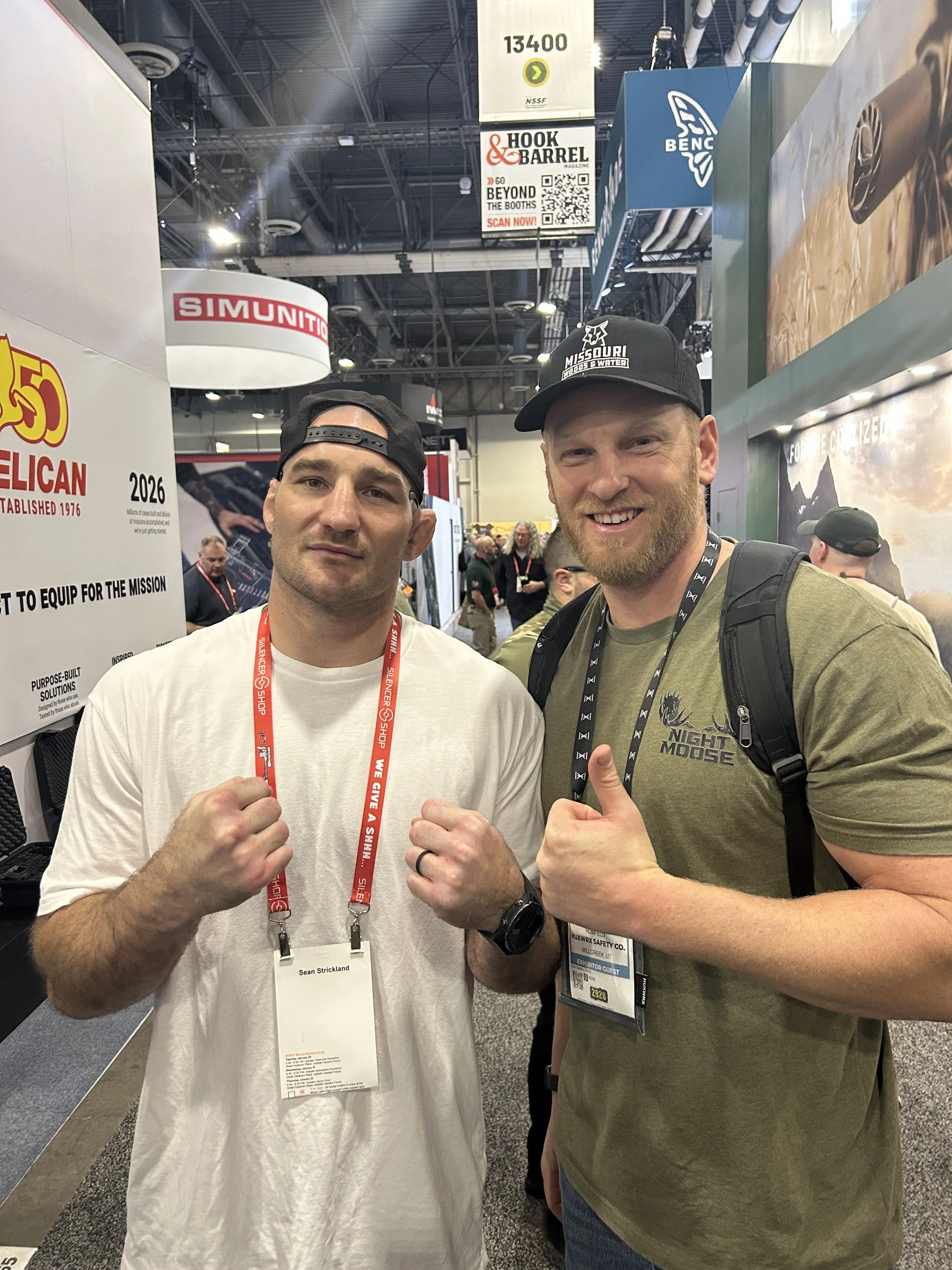 Two men standing at a trade show, both displaying a fist bump and smiling. The man on the left wears a white T-shirt and a backward black cap, has a red lanyard with a name tag, and makes a fist gesture. The man on the right wears a green T-shirt wit