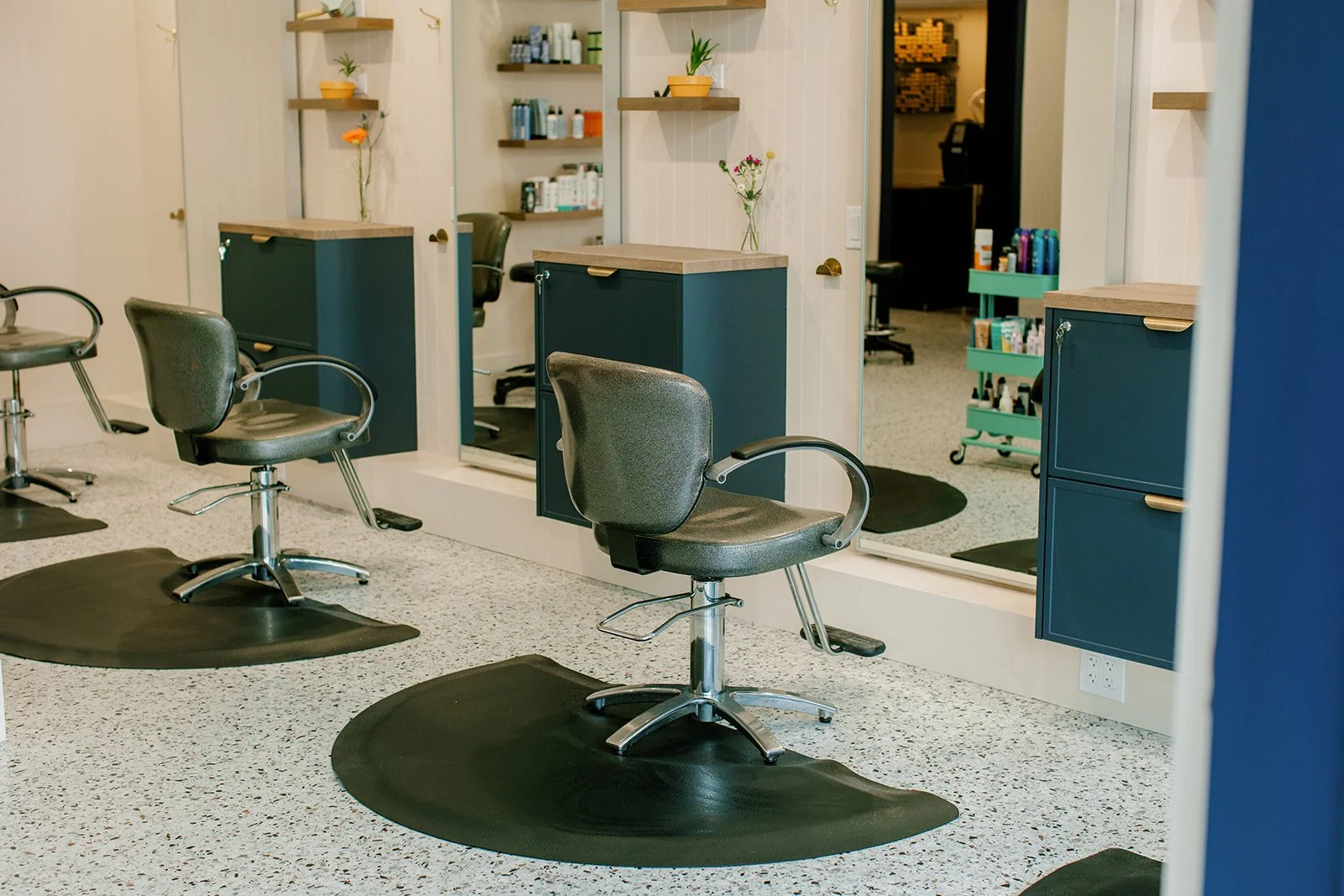 B Parlor, a modern hair salon with black swivel chairs that can hold up to 400lb. , large mirrors, and shelves with hair products, decorated with flowers in vases.