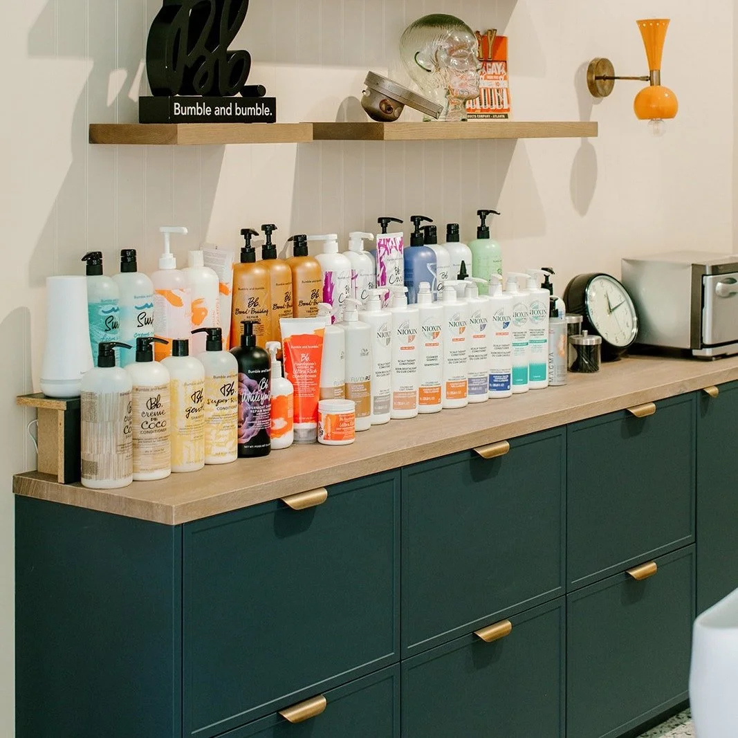 B Parlor Salon's backbar area with bottles of hair and skincare products, towel warmer, and decorative items on a wall shelf.