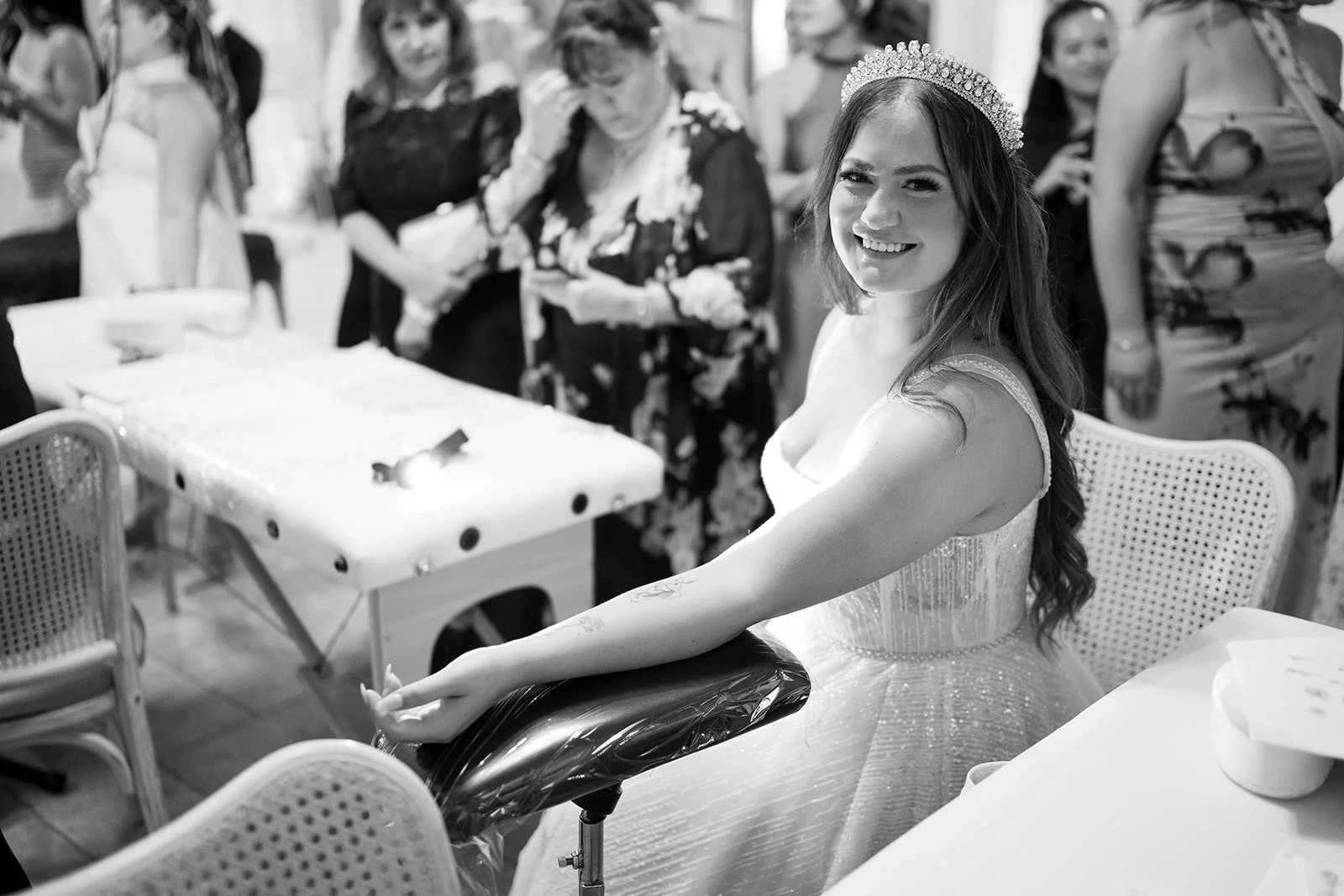 A woman wearing a wedding dress and tiara is smiling while sitting at a table, with her arm extended showing a tattoo, during a celebration with other women in the background.