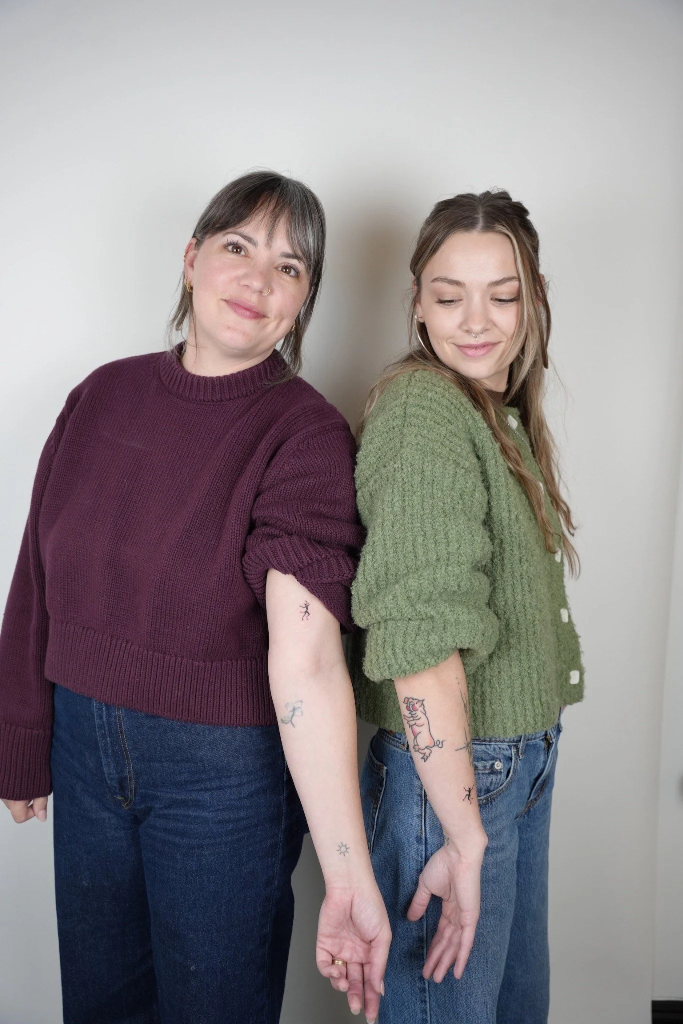 Two women stand side by side against a plain wall. The woman on the left has short, dark hair with bangs and is wearing a maroon sweater. The woman on the right has long, light brown hair and is wearing a green cardigan. Both women have tattoos on th
