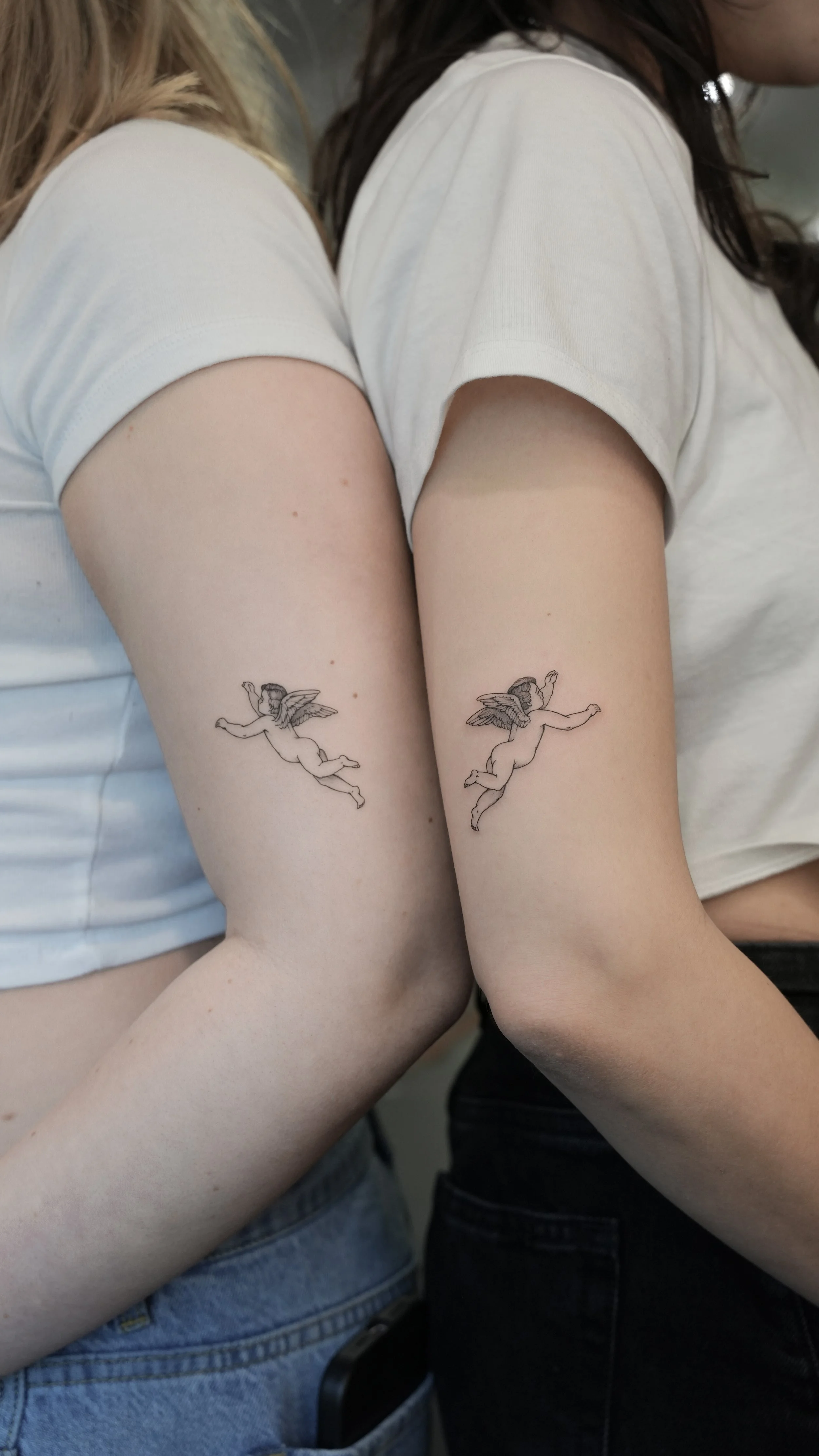 Two women standing back to back, each with a tattoo of a flying cherub on her upper arm.