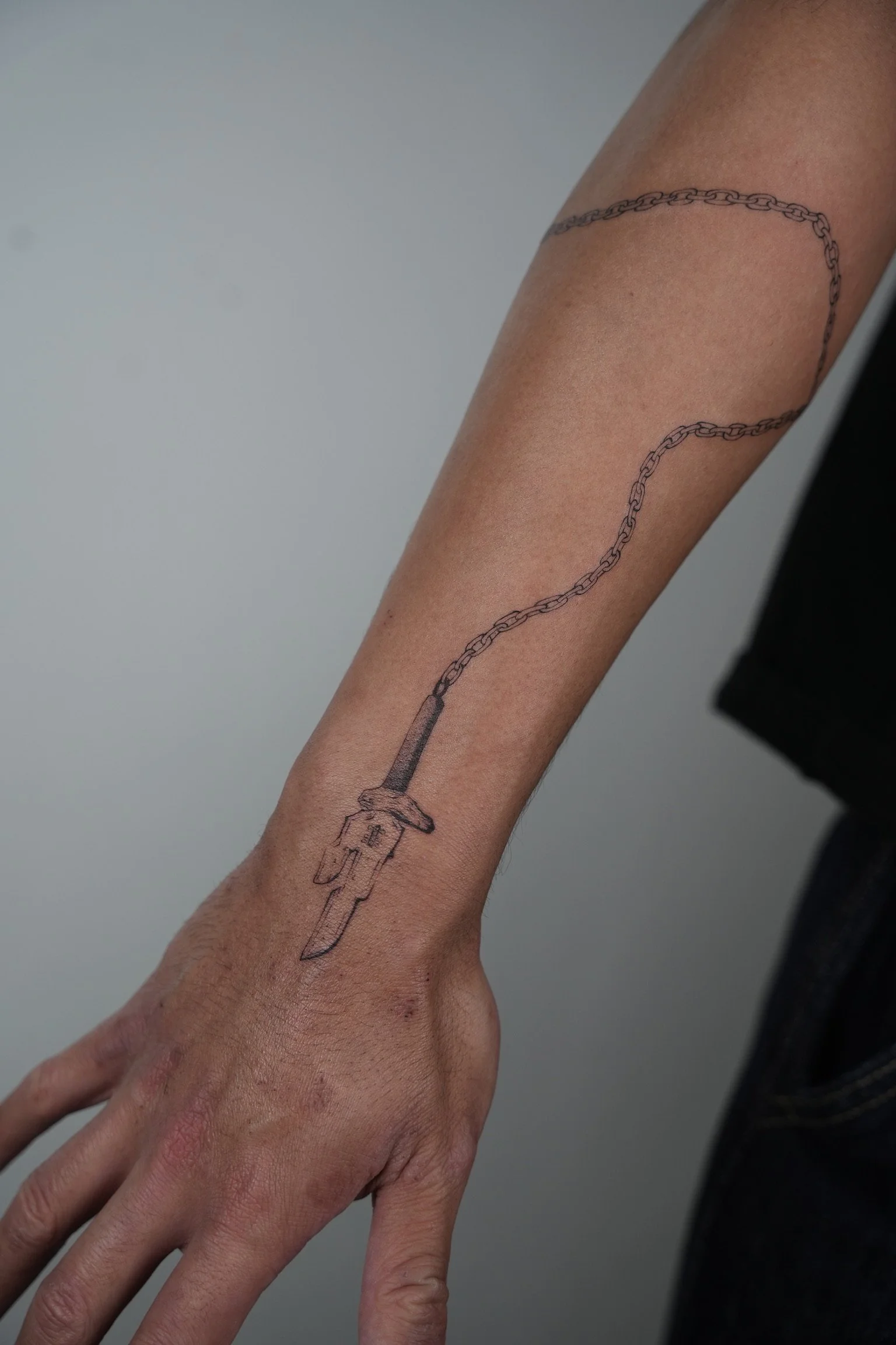 Tattoo of a syringe with a chain extending from its barrel on a person's forearm.