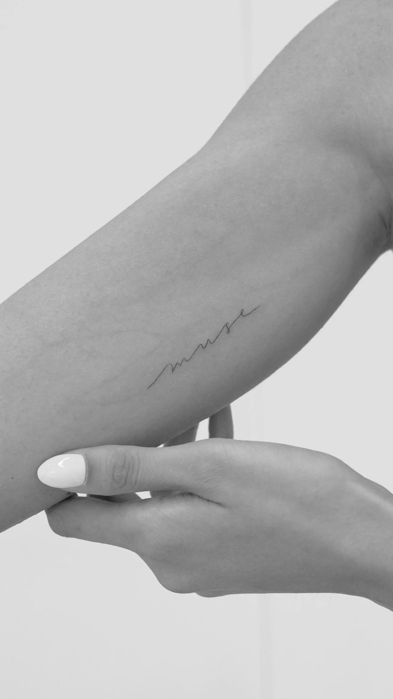 Close-up of a person's hand holding their arm, showing a black script tattoo on the inner wrist area. The tattoo appears to be a word written in cursive.