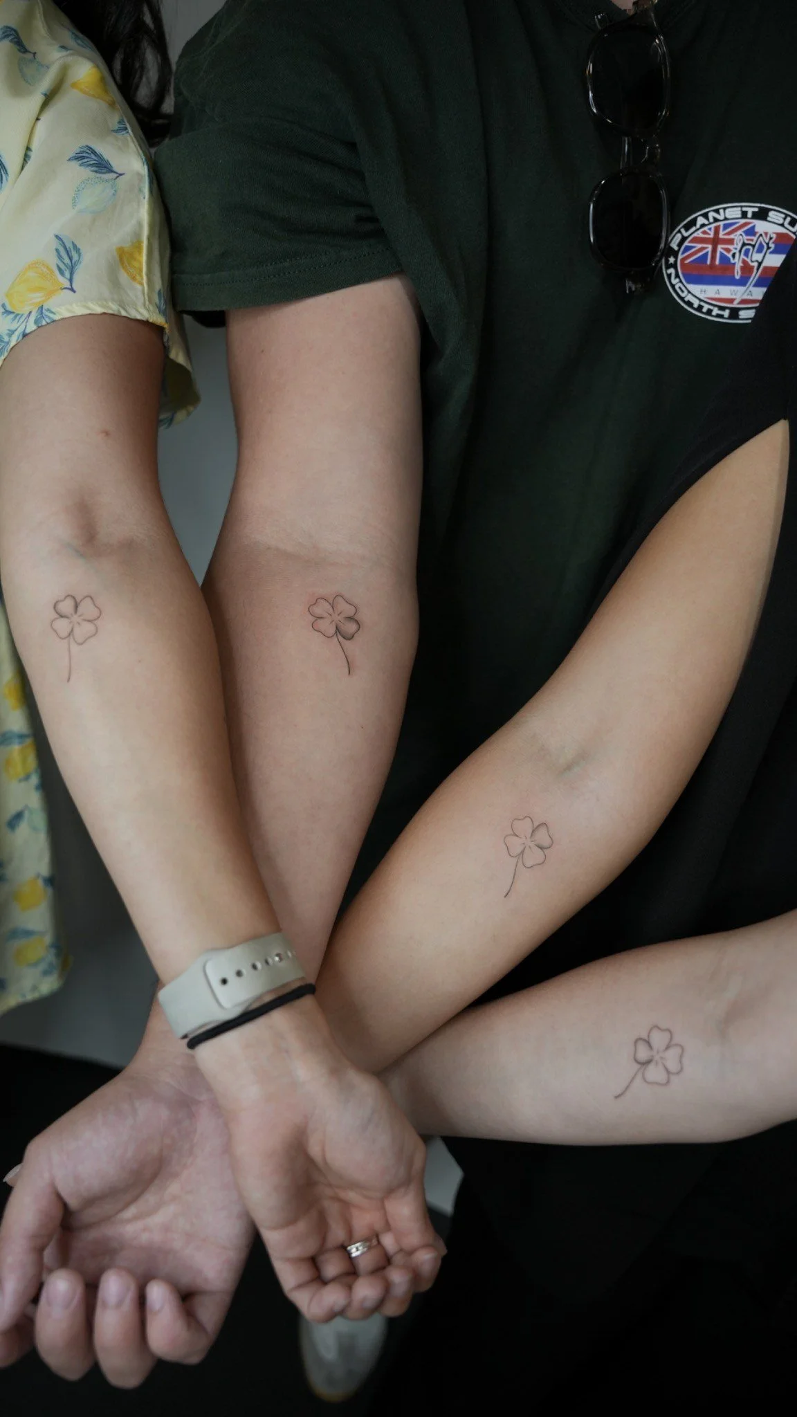 Close-up of four arms with small, simple four-leaf clover tattoos, intertwined and holding hands, showing affection or friendship.
