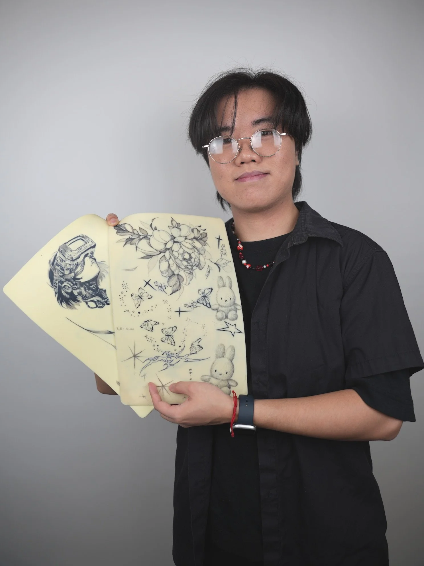 A person with glasses and a black shirt holding up paper sheets with sketches of a girl with goggles, flowers, butterflies, stars, and rabbits against a plain gray background.