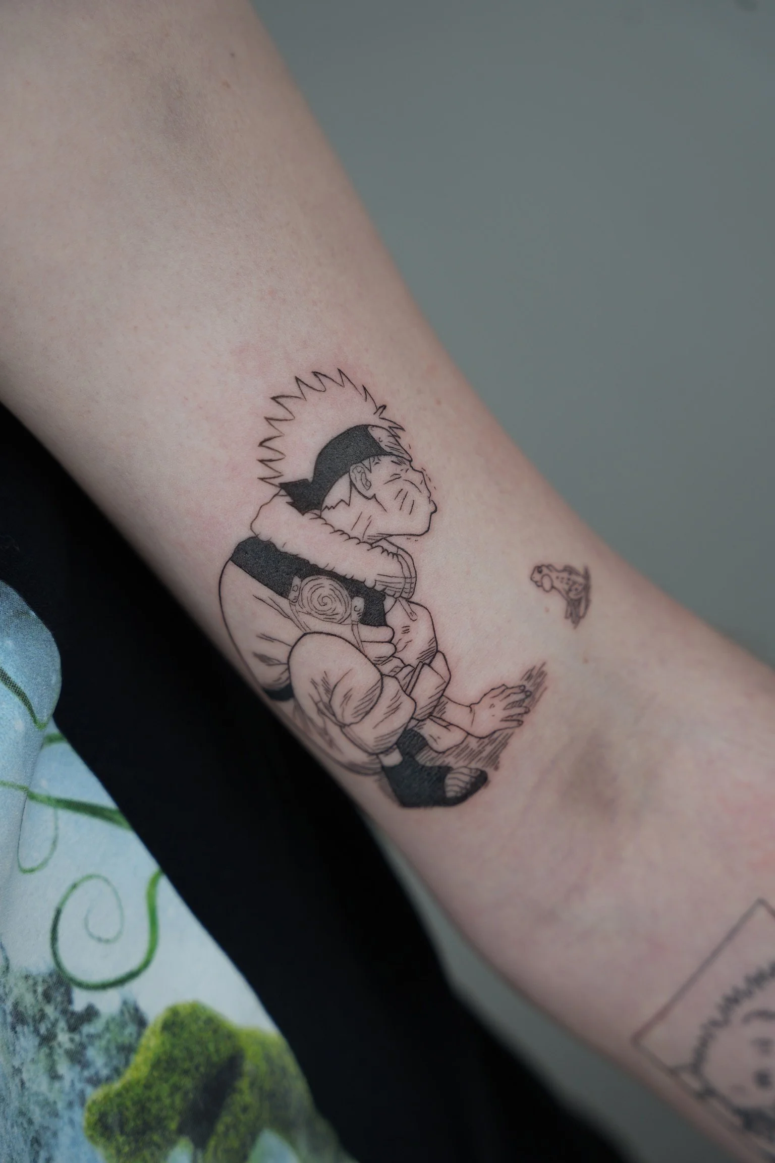 Black and gray tattoo of a young boy with spiky hair, face markings, a headband, and a scarf, sitting with eyes closed, next to a small flying robot or drone.