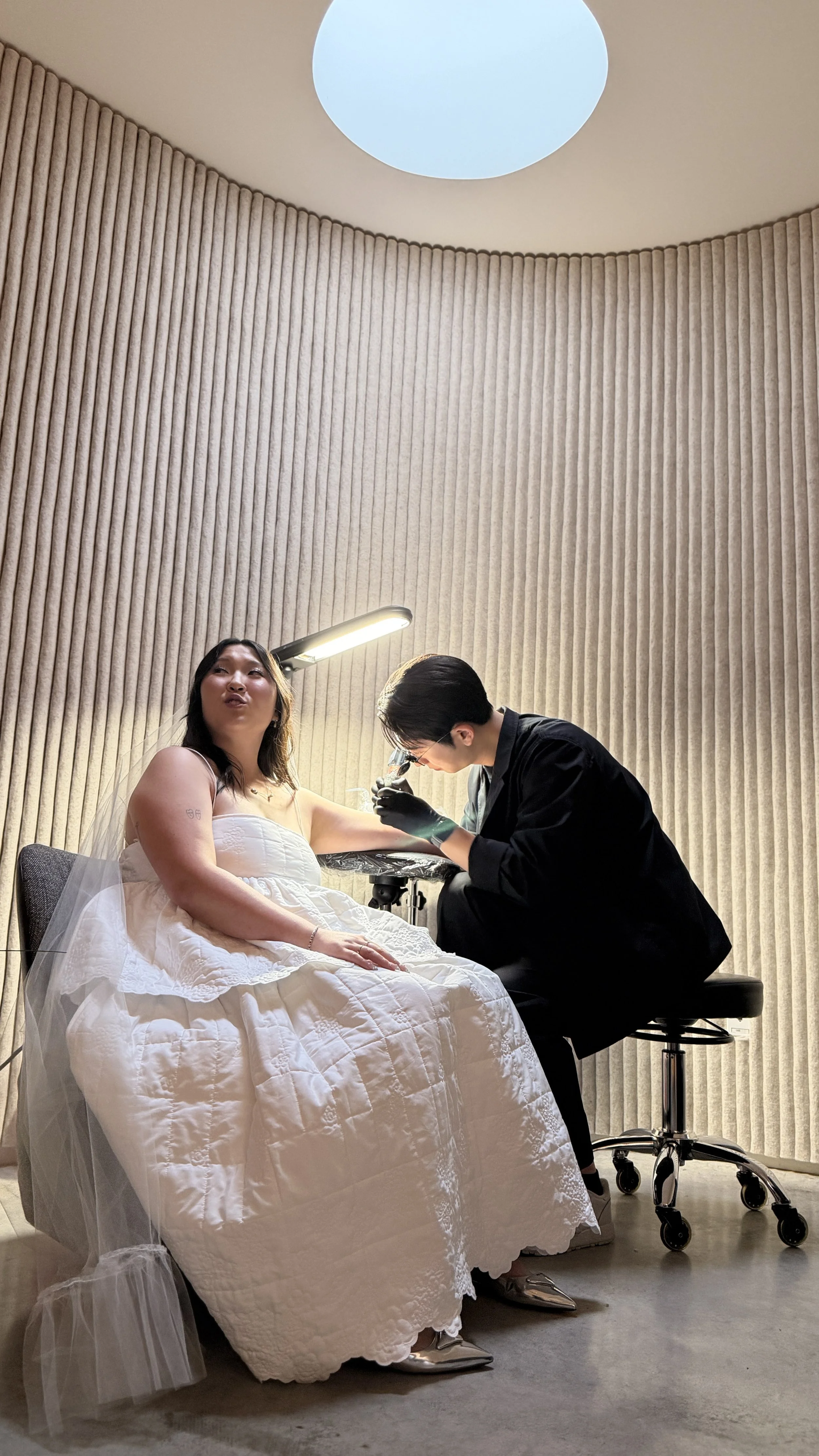 A woman in a white dress sitting on a bed getting a tattoo on her arm, while a tattoo artist in black clothing and gloves works on her arm in a room with beige curtains and a ceiling light.