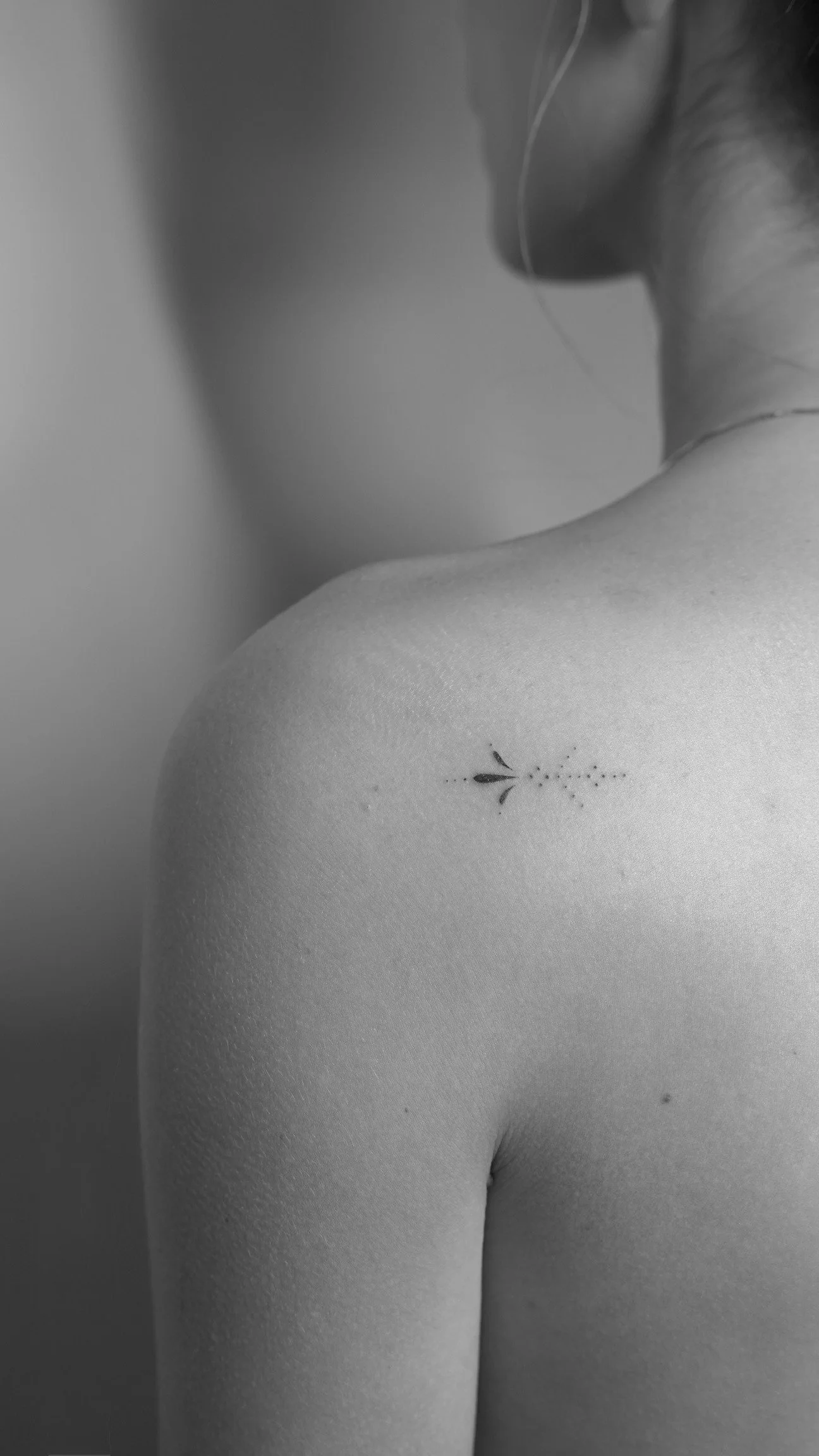 Close-up black-and-white photograph of a woman's shoulder and back, showing a small tattoo of a simple plant with leaves and dots.