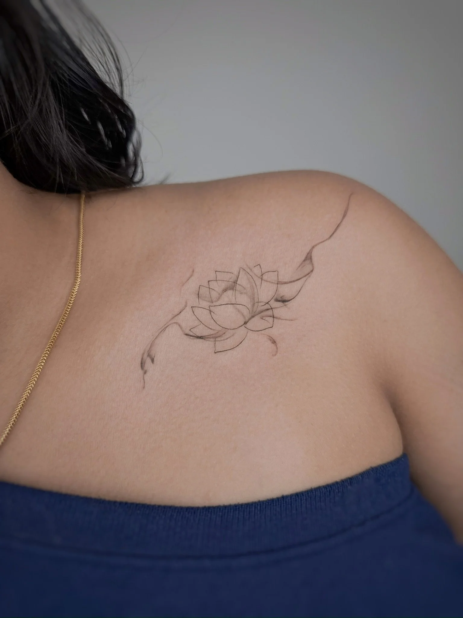 Close-up of a shoulder with a delicate lotus flower tattoo and a gold chain necklace, partially visible, wearing a blue strapless top.