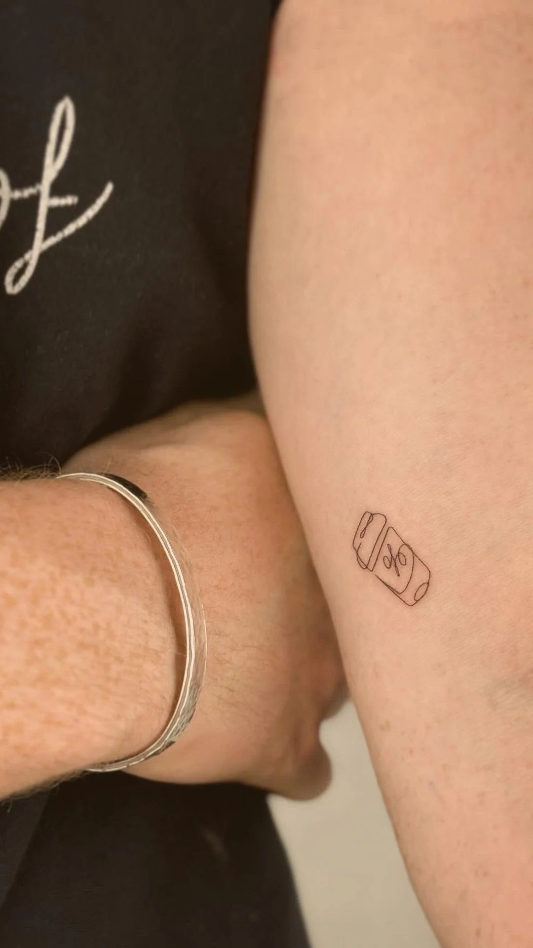 A close-up of two arms, one wearing a thin silver bracelet and the other with a small tattoo of a battery with a percentage sign, and a black shirt with white script partially visible.