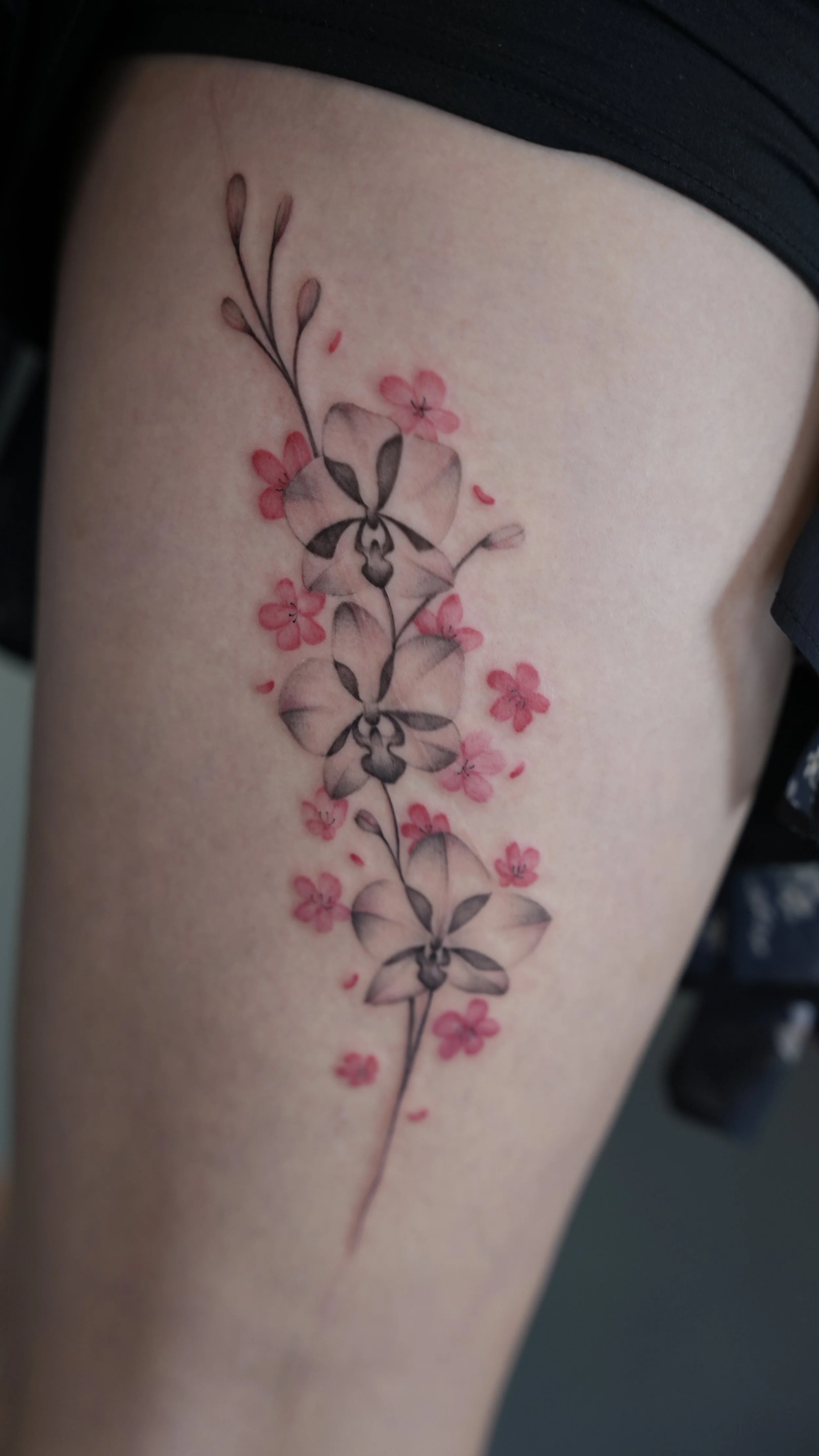 A floral tattoo featuring black and gray orchids with pink flowers and petals on a person's thigh.
