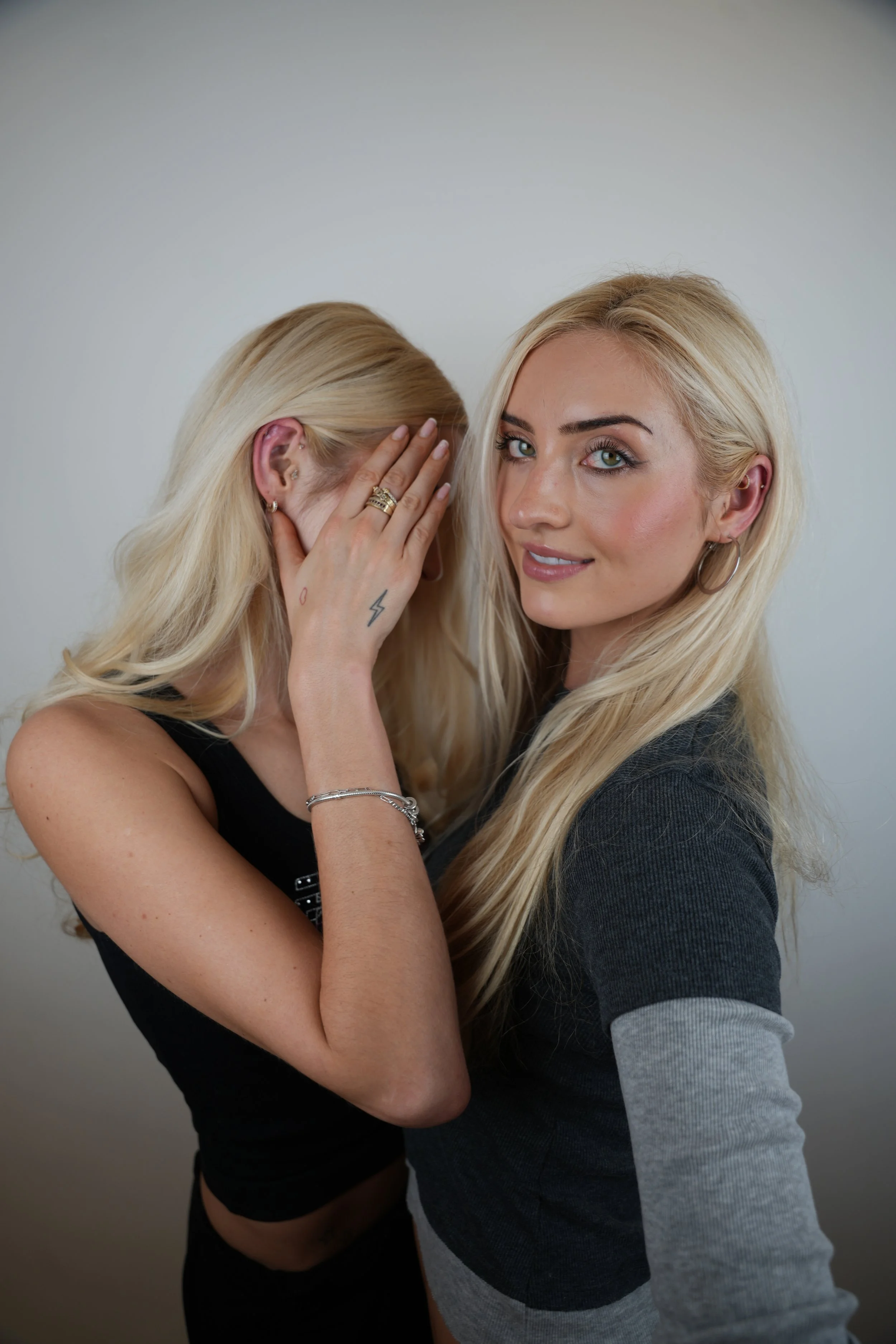 Two young women with blonde hair, one is smiling and looking at the camera, the other has her face covered with her hand. They are standing against a plain background and showcasing new piercing that they just got from Matching Piercing Deals in NYC