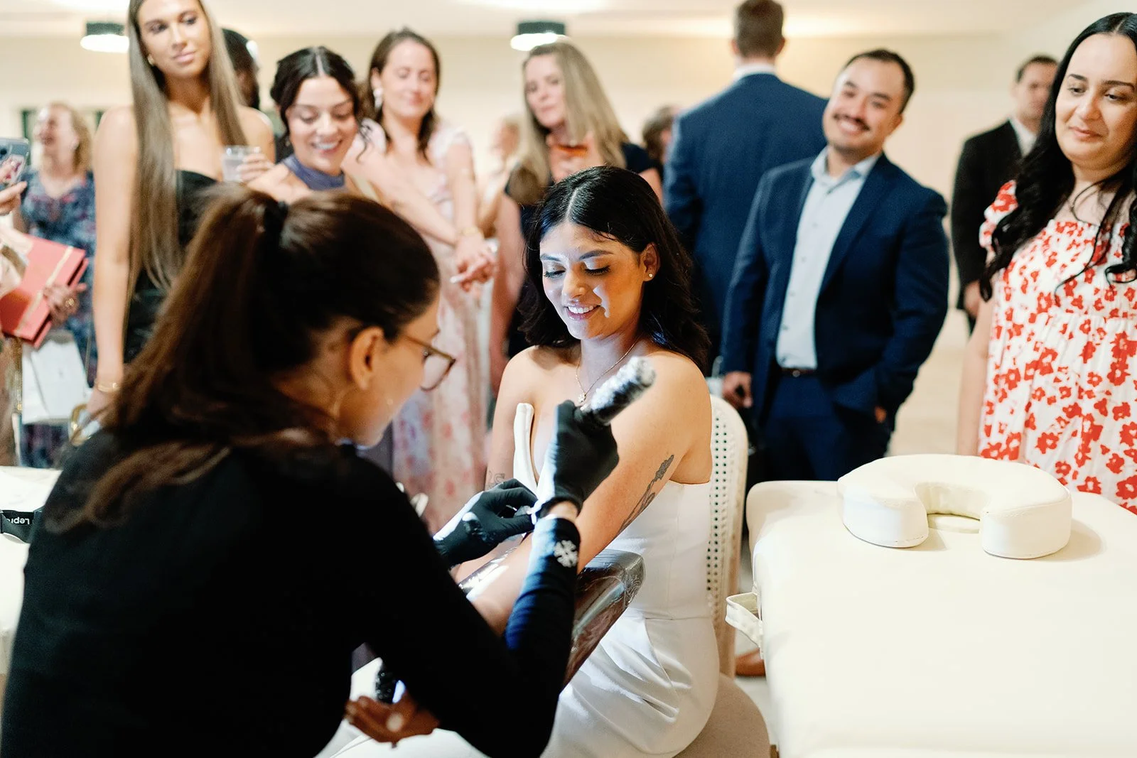A woman in a white dress gets a tattoo on her arm at a tattoo event, surrounded by smiling onlookers in formal attire.
