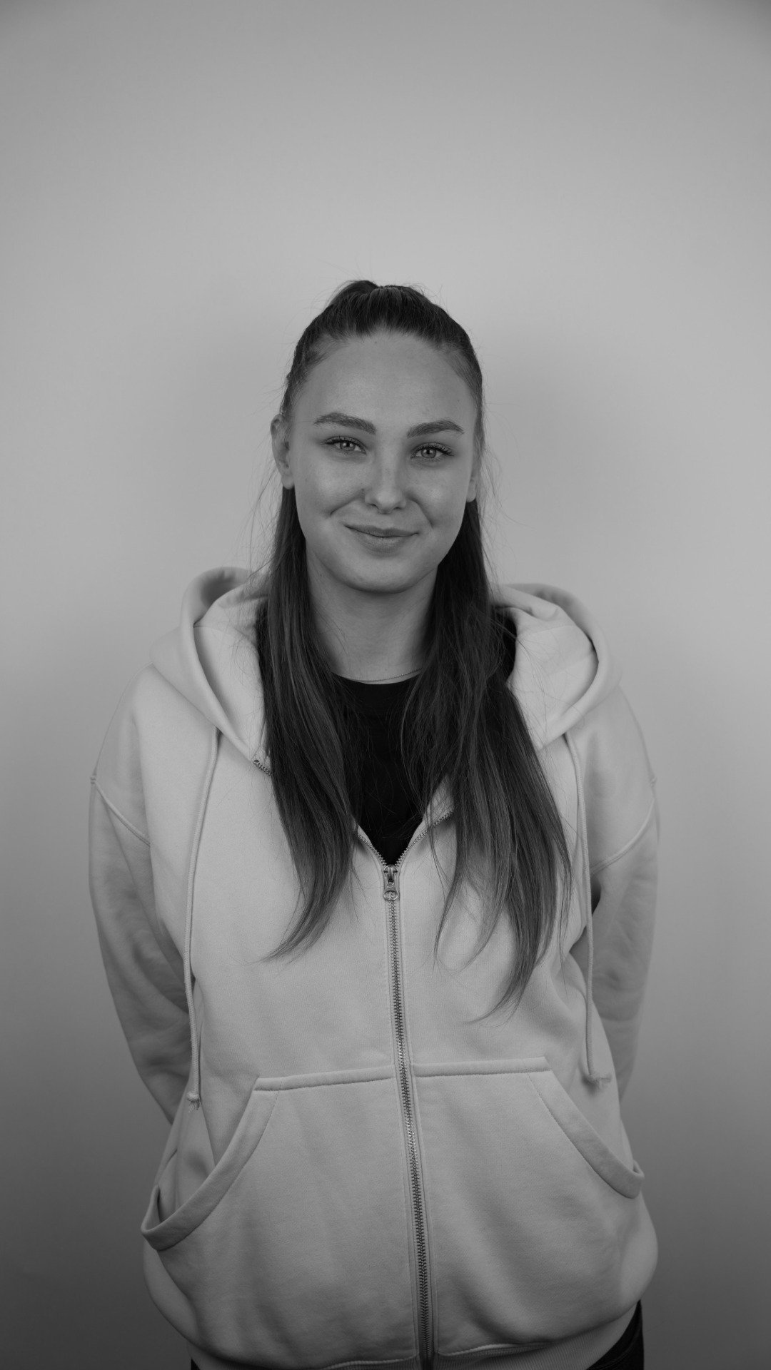 Black and white photo of a young woman with long hair in a hoodie, smiling, standing against a plain background.