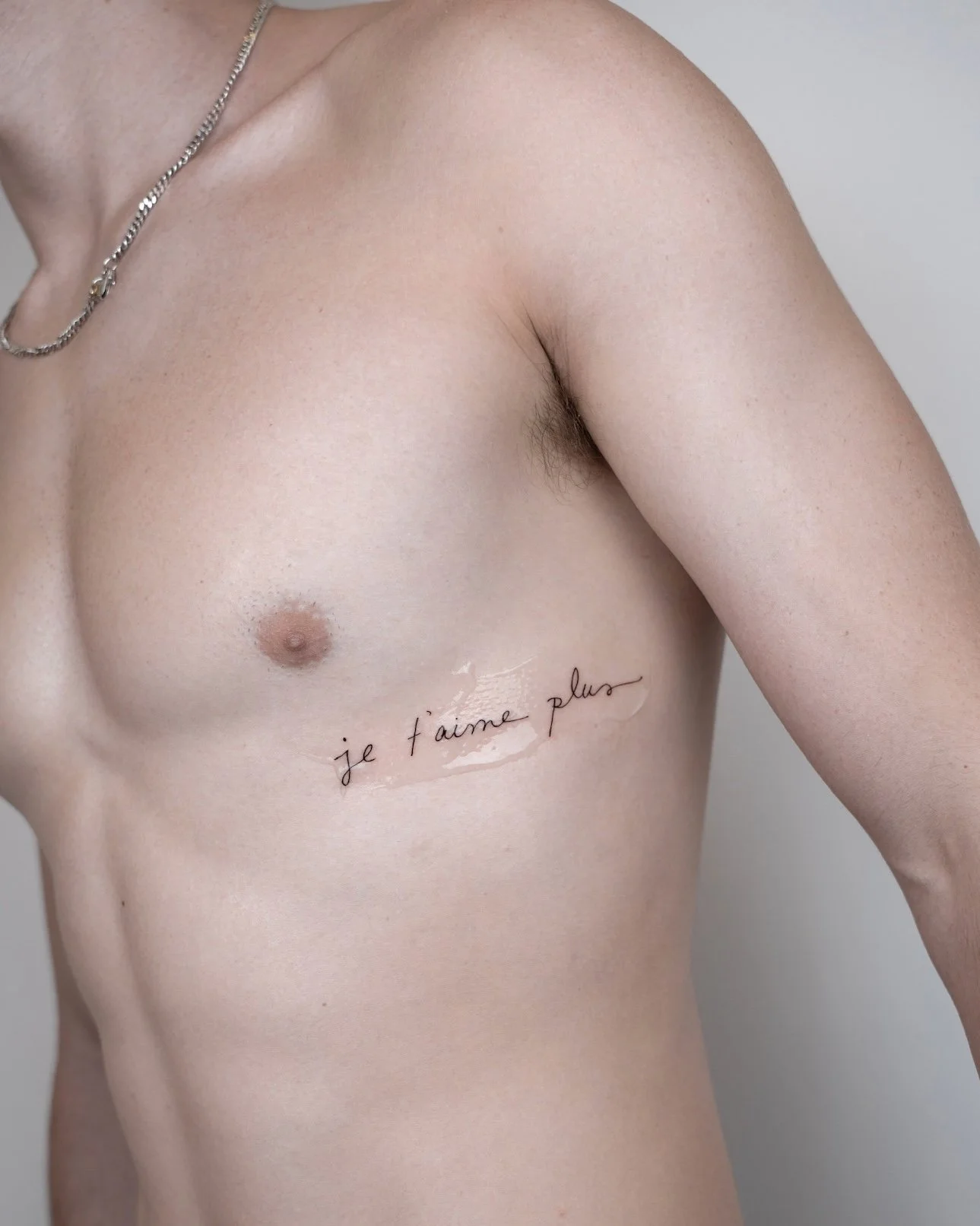 A close-up of a person's upper torso, showing the chest and shoulder, with a tattoo of French text saying 'je t'aime plus' across the ribcage.
