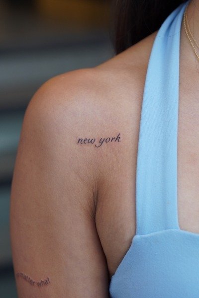 Close-up of a woman's shoulder with a tattoo that says "new york" and part of a light blue top.