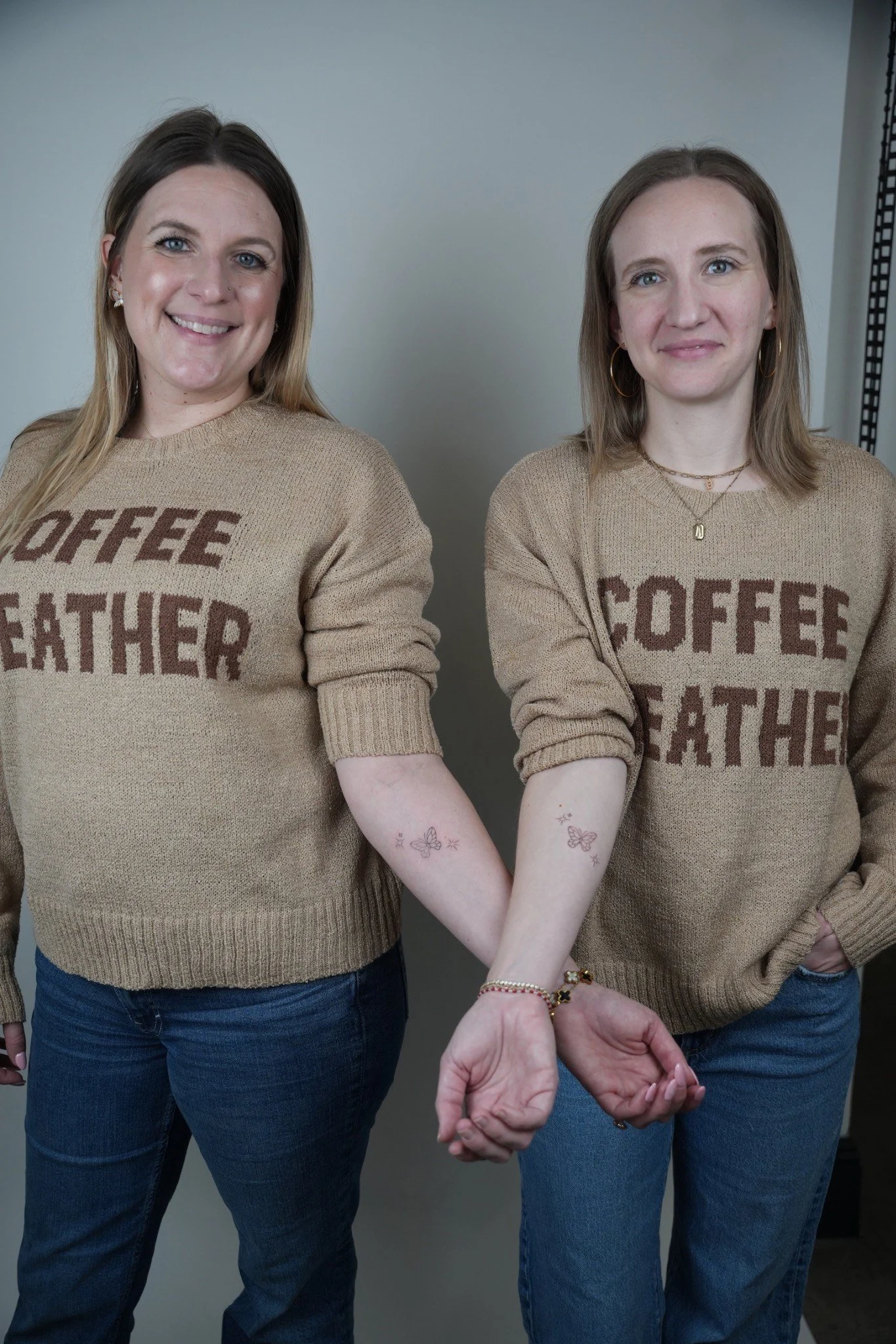 Two women holding hands, wearing matching beige sweaters with the words 'coffee' and 'weather' knitted into them, standing side by side in front of a light gray wall.