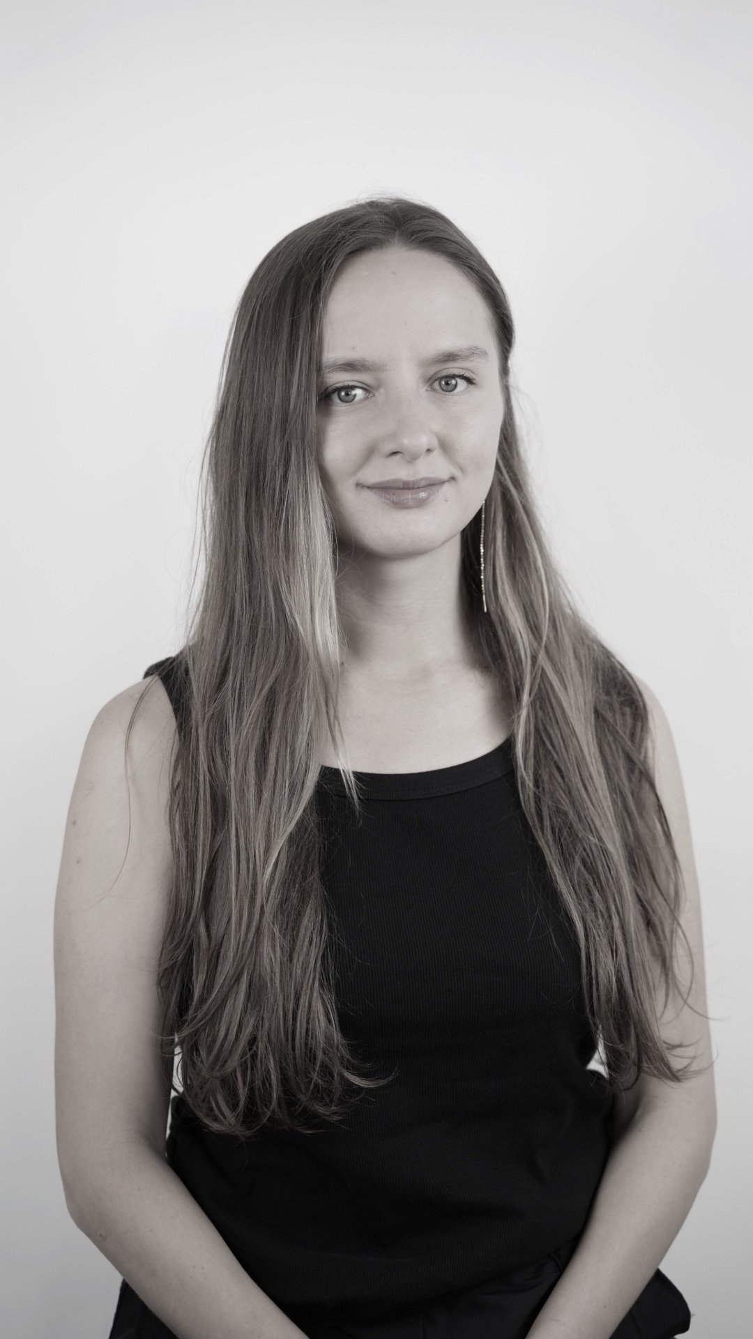 A woman with long wavy hair wearing a sleeveless black top, standing against a plain white background.