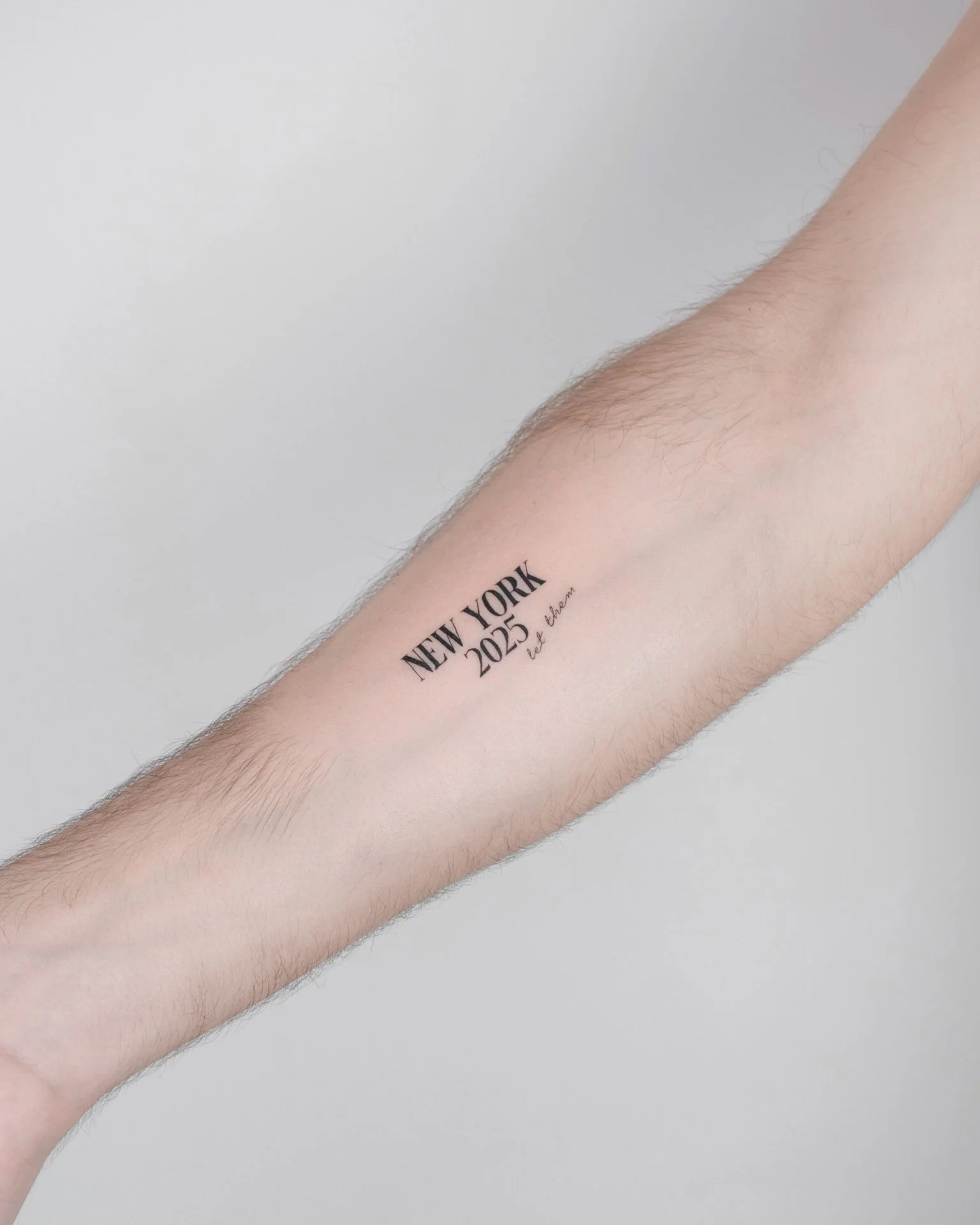 Tattoo on person's forearm reading "NEW YORK 2025 let them in" with minimal hair on arm and a plain light background.