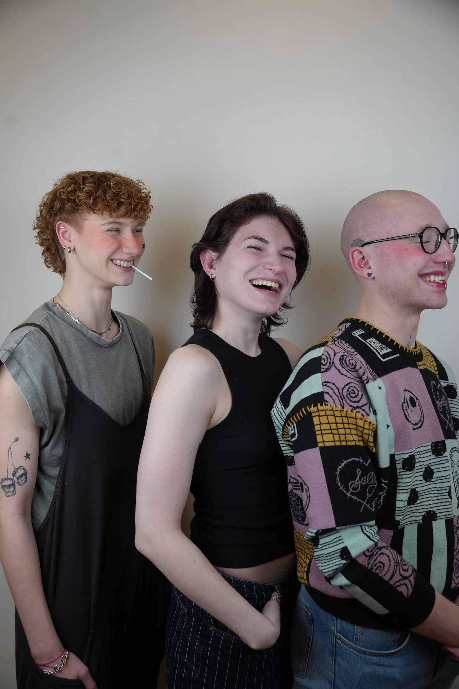 Three young adults smiling and enjoying themselves, standing in a line against a plain wall.