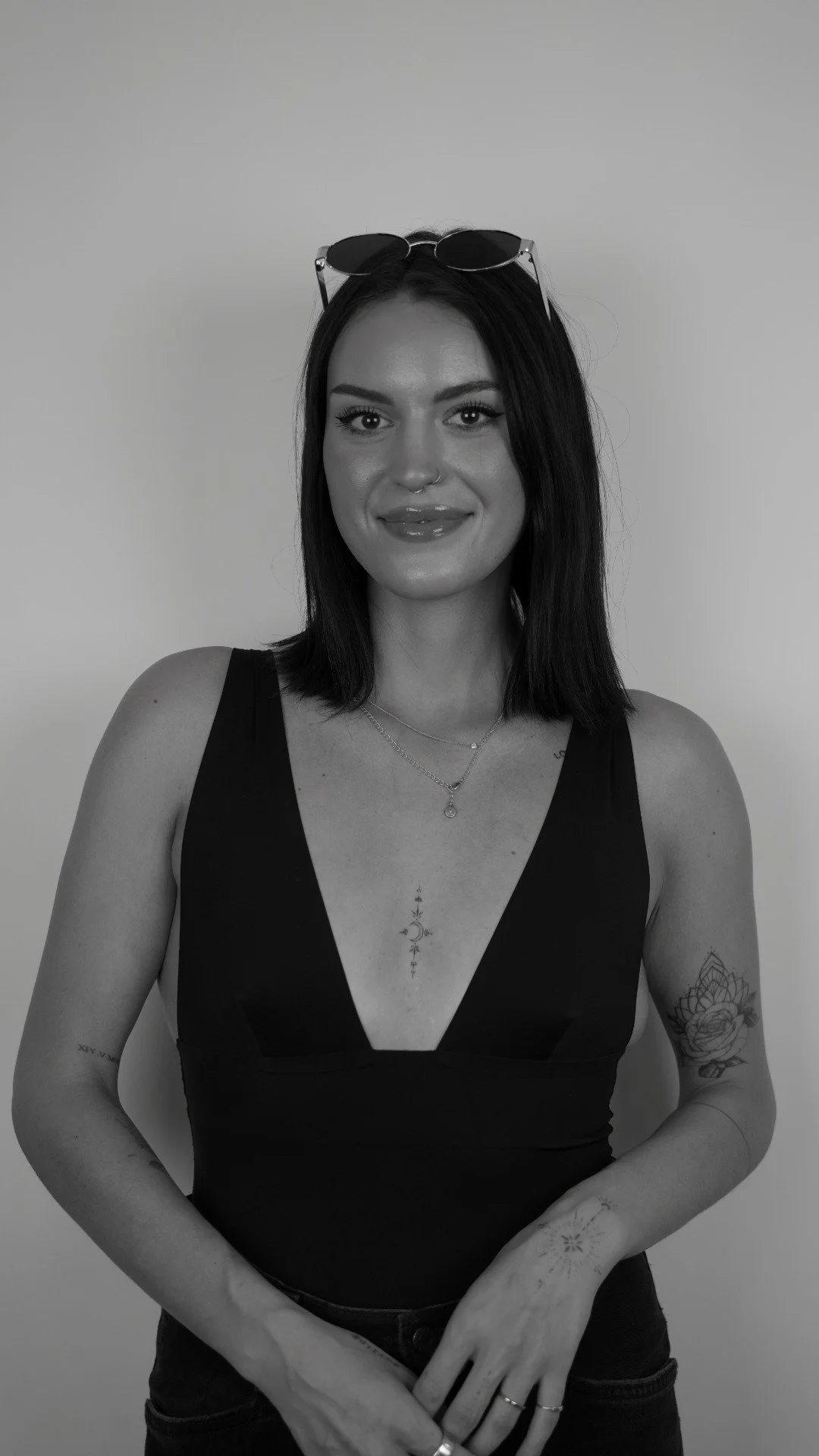 A woman with long dark hair wearing sunglasses on her head, a black tank top, and layered necklaces, smiling at the camera, standing against a plain wall, with tattoos on her left arm and chest.