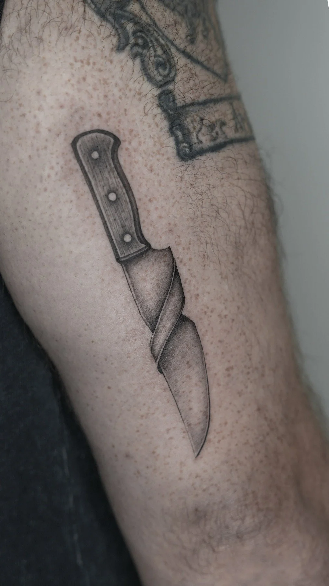A tattoo of a kitchen knife with a detailed handle and a sharp, pointed blade on a person's upper arm.