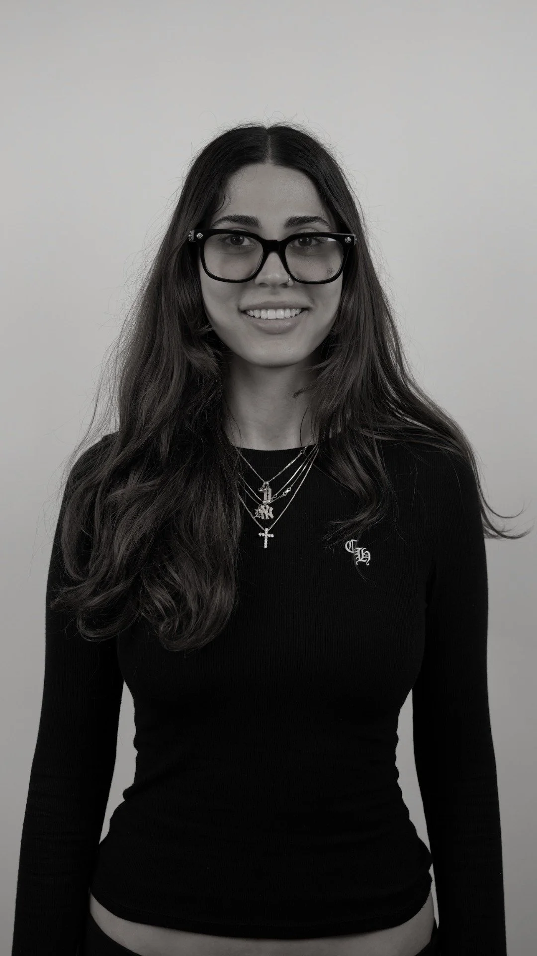 A young woman with long wavy hair, black glasses, and layered necklaces, smiling in a black long-sleeve shirt against a plain background.