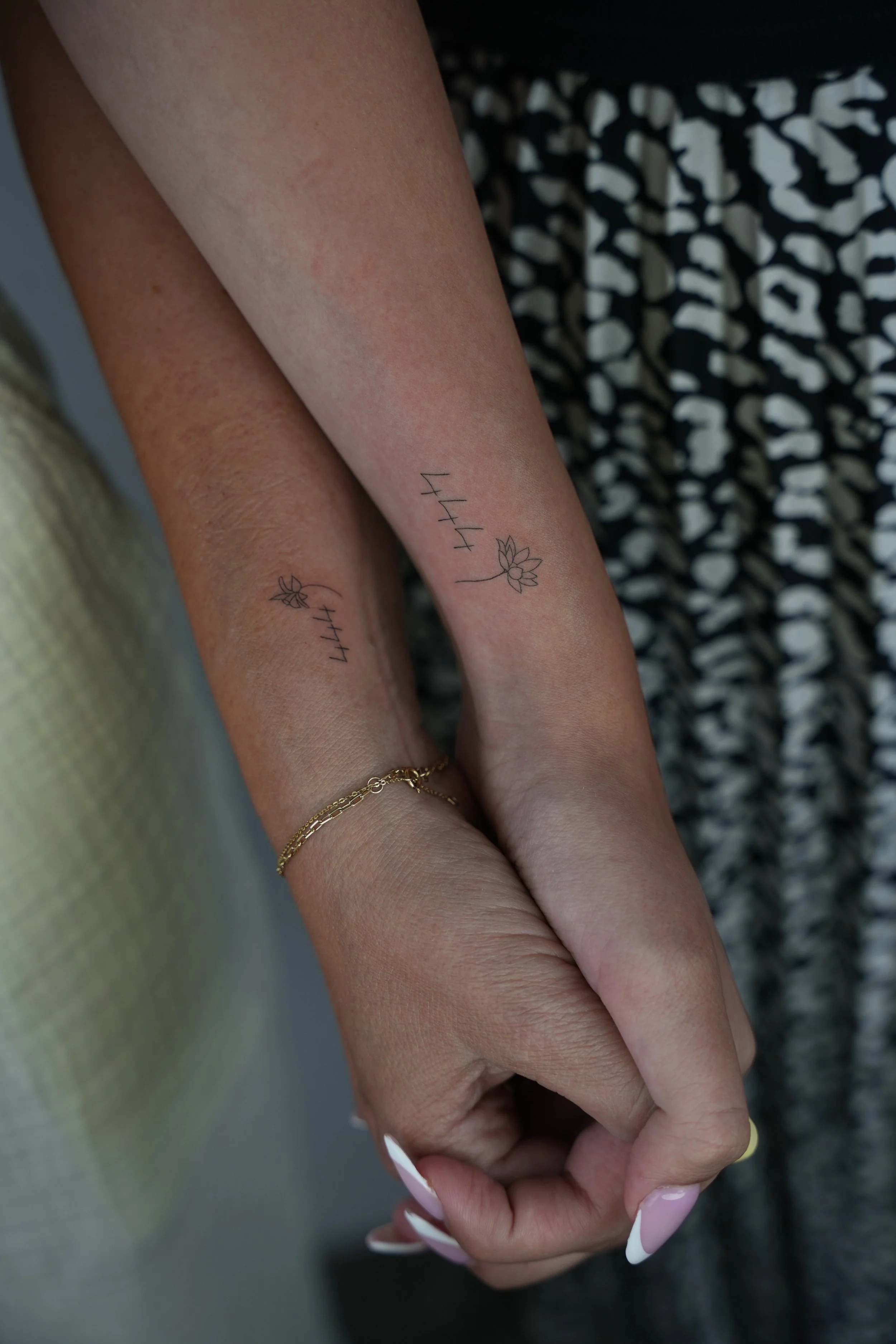 Two arms with matching tattoos of the number '444' and a lotus flower. One arm has a gold bracelet. The background shows part of a patterned dress.