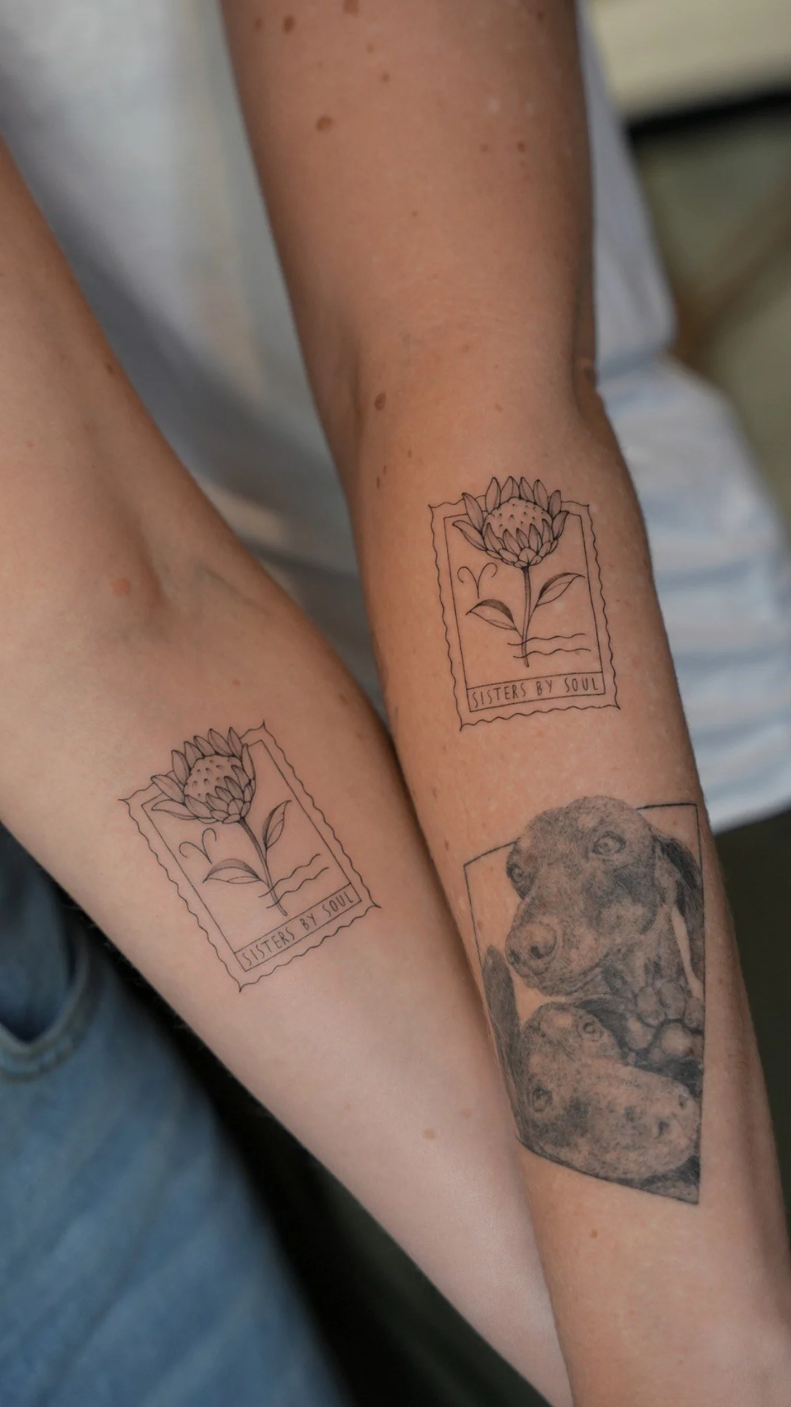 Two people with matching tattoo designs on their forearms, featuring a flower inside a rectangular frame with the text 'SISTERS BY SOUL'.
