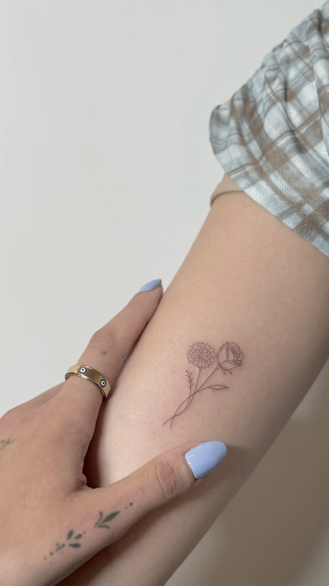 Close-up of a person's arm with a black floral tattoo, showing a dahlia and roses, with their hand gently touching the arm. The person has light blue painted nails and is wearing gold rings. Part of their plaid shirt is visible.
