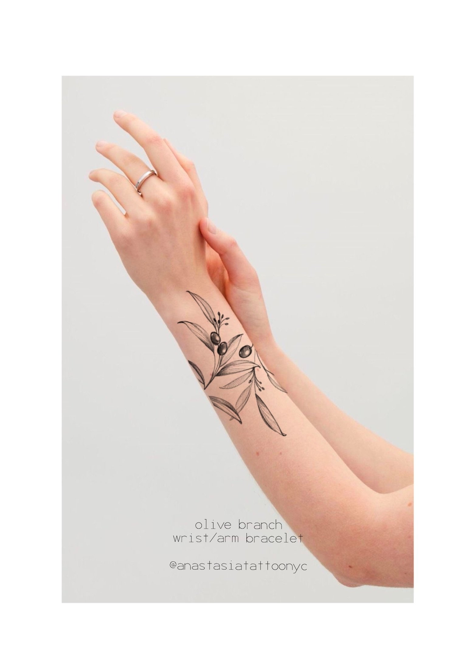 A person's left forearm with a black ink tattoo of olive branches and leaves. The person is wearing two silver rings on their fingers and is holding their right hand with their left hand. The background is plain light gray. Text at the bottom of the 