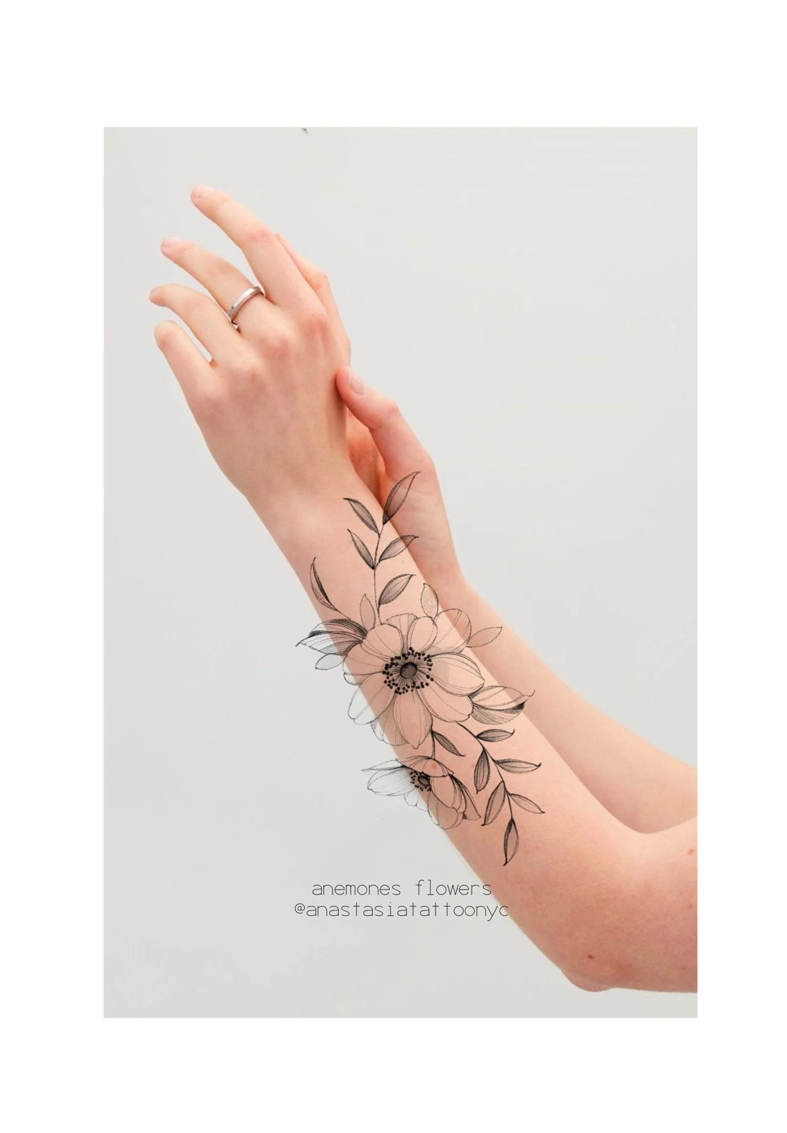A person's arm with a floral tattoo design of anemone flowers and leaves, along with a finger ring, against a plain background. Text indicates the tattoo is by Anastasia Tatt Too NYC.