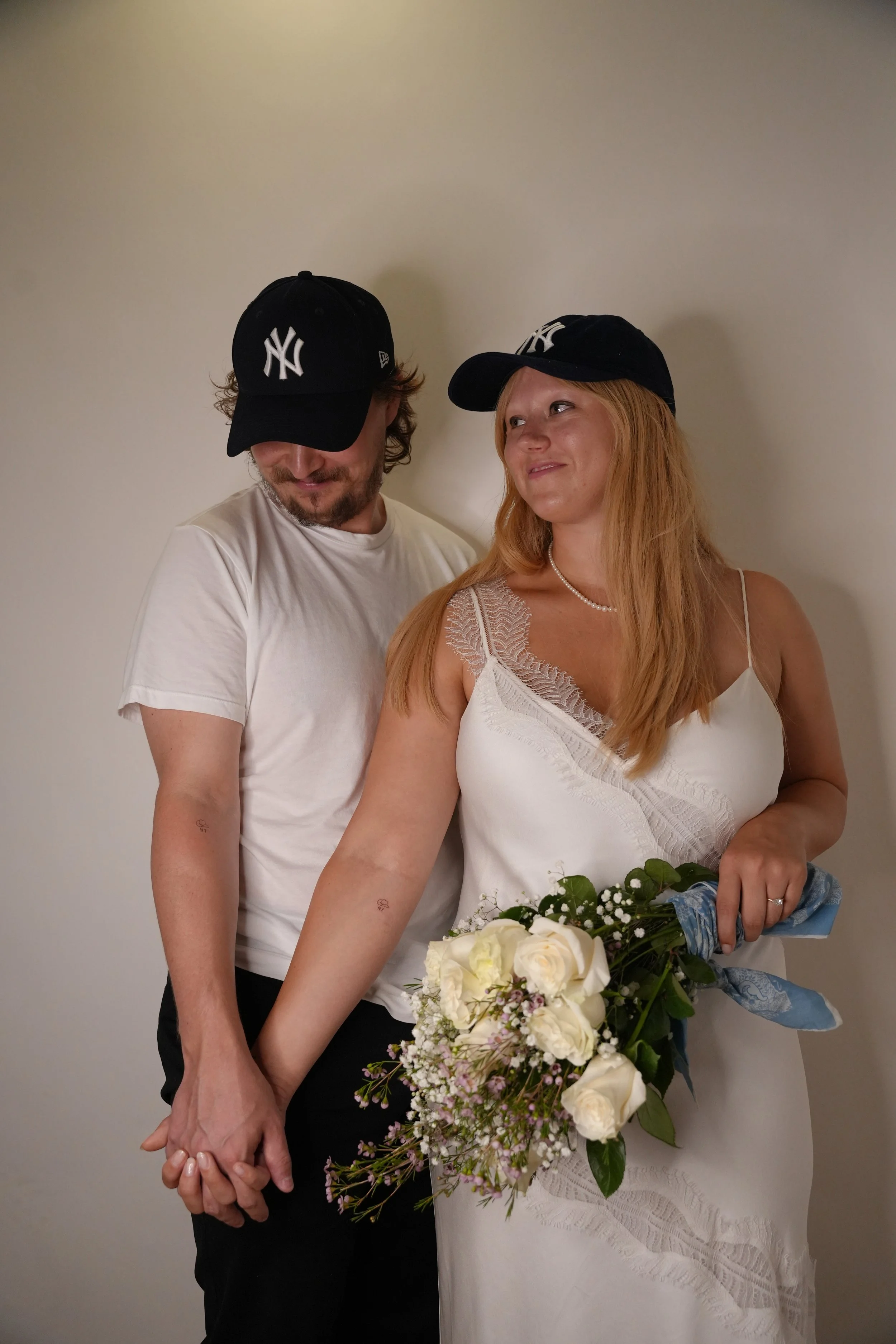 A couple holding hands, with the woman holding a bouquet of white roses, standing against a plain wall. Both are wearing black New York Yankees caps and casual clothing.