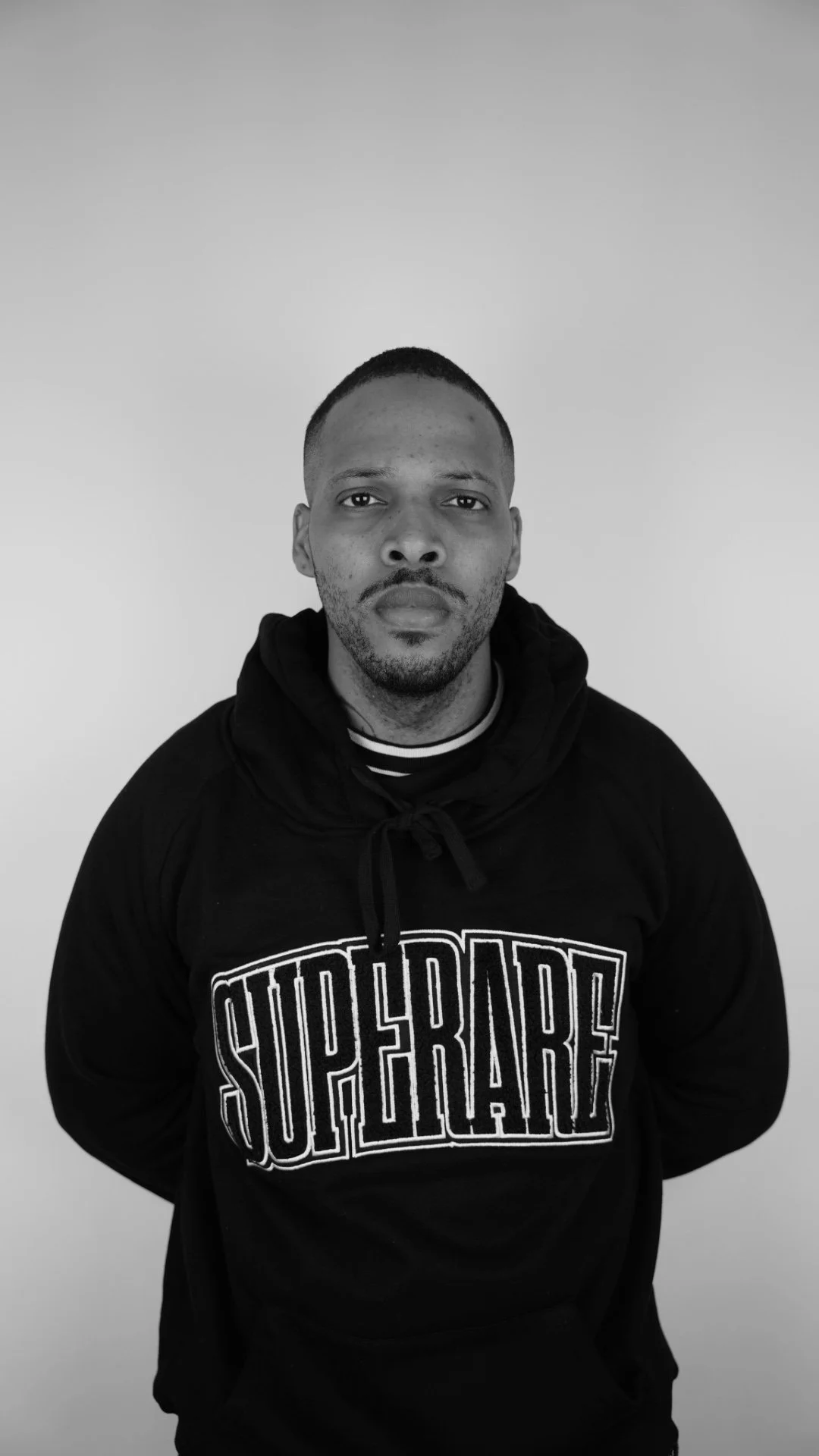 Black and white portrait of a man with short hair and facial hair, wearing a hoodie with 'SuperRad' written on it, against a plain background.