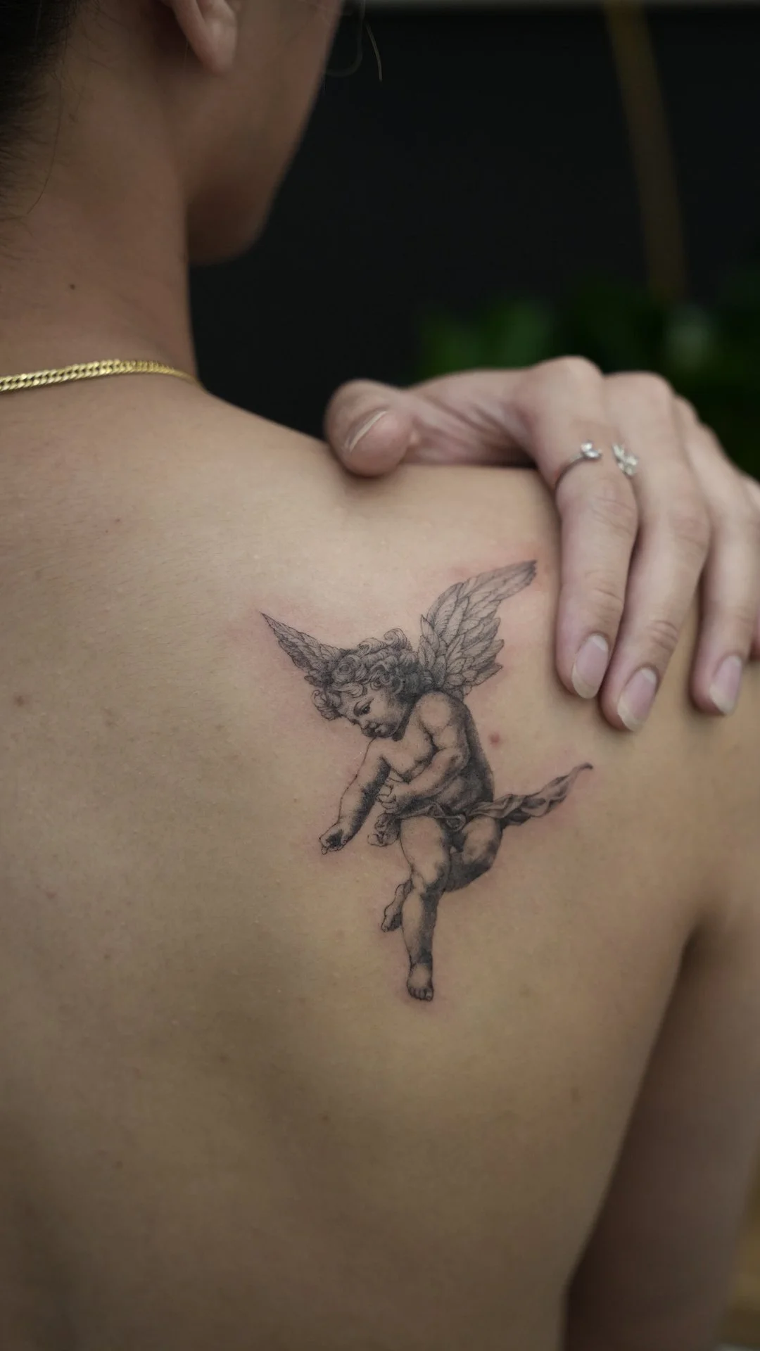 A tattoo of a cherub with wings, sitting and reading a book, is on a person's shoulder.