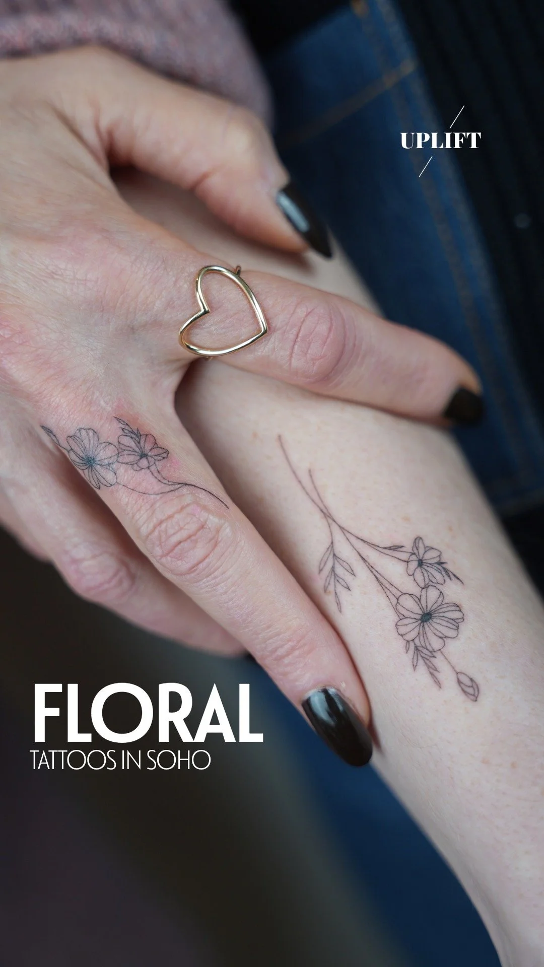 Close-up of a hand with black nail polish, holding another person’s arm, showing floral tattoos. A gold heart-shaped ring is on the finger. The tattoos are delicate line art of flowers and leaves.