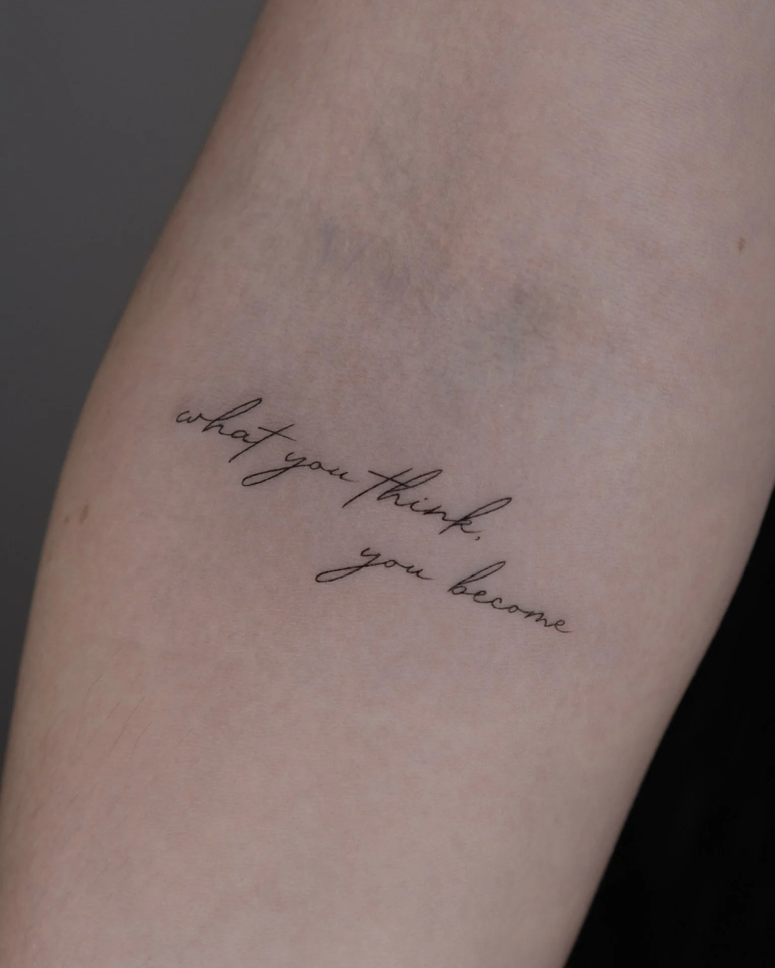 Close-up of a forearm with a tattoo that reads, 'what you think, you become' in cursive black ink.