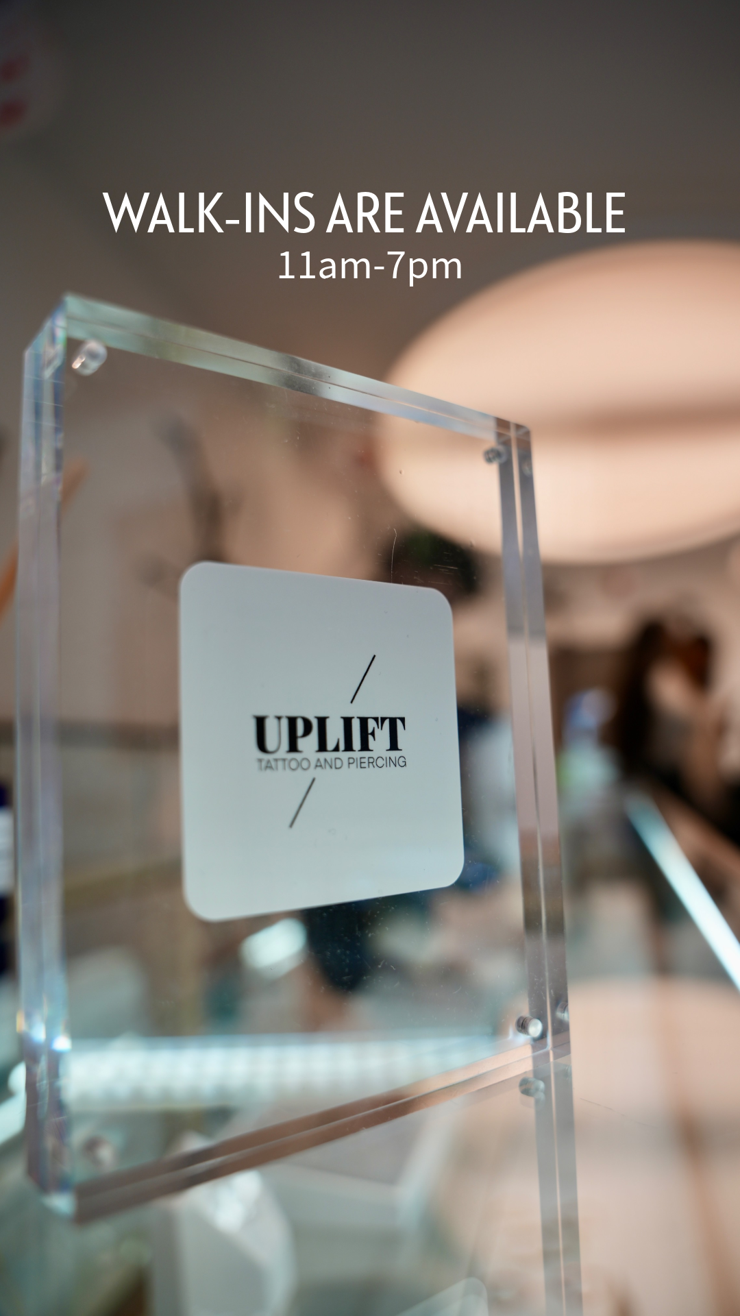 Sign on a clear acrylic stand announcing walk-in availability for UPLIFT tattoo and piercing from 11am to 7pm.