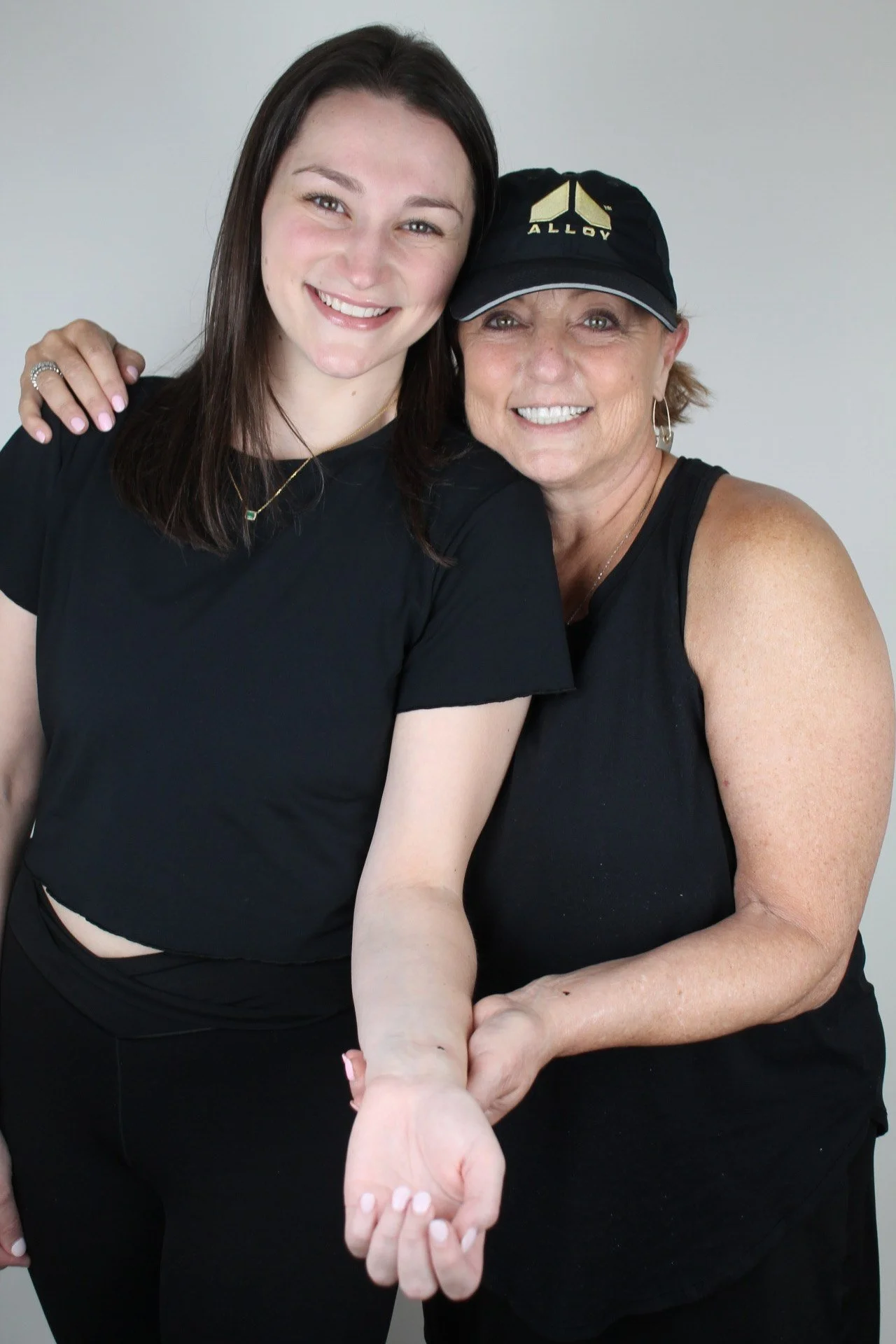 Two smiling women, one younger and one older, taking a selfie together. The older woman is wearing a black cap and bracelet, and the younger woman is wearing a black shirt and has her hand on the older woman's shoulder.