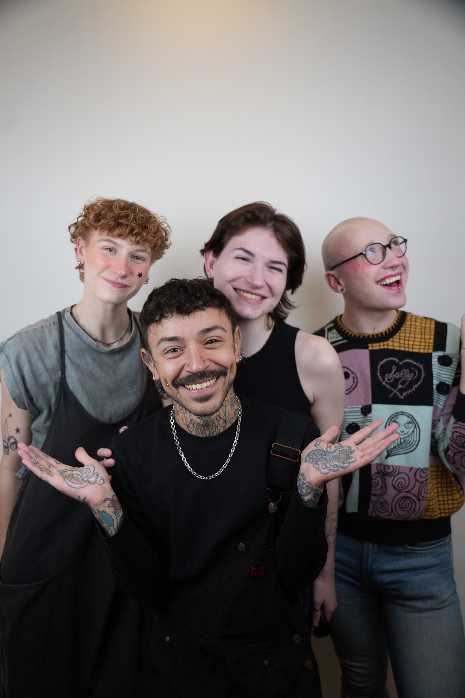 Four diverse young adults smiling and posing in front of a plain wall, with one person in the front with tattoos and a necklace, and three others behind them, one with a star on the cheek, another with short brown hair, and the last with glasses and 